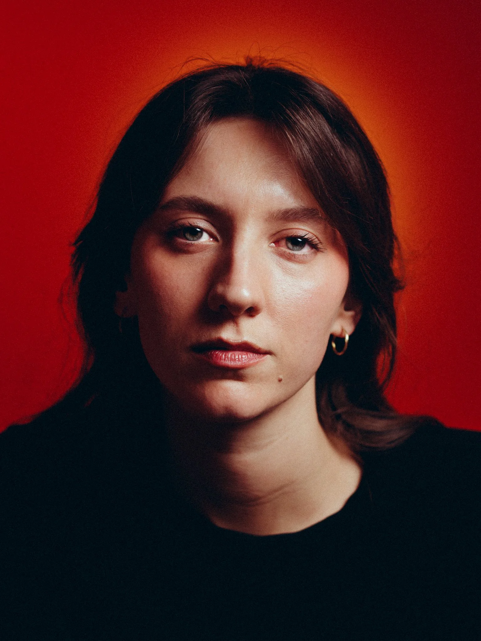 Close-up portrait of a woman with dark hair and blue eyes, wearing a black top and small hoop earrings, against a red background.