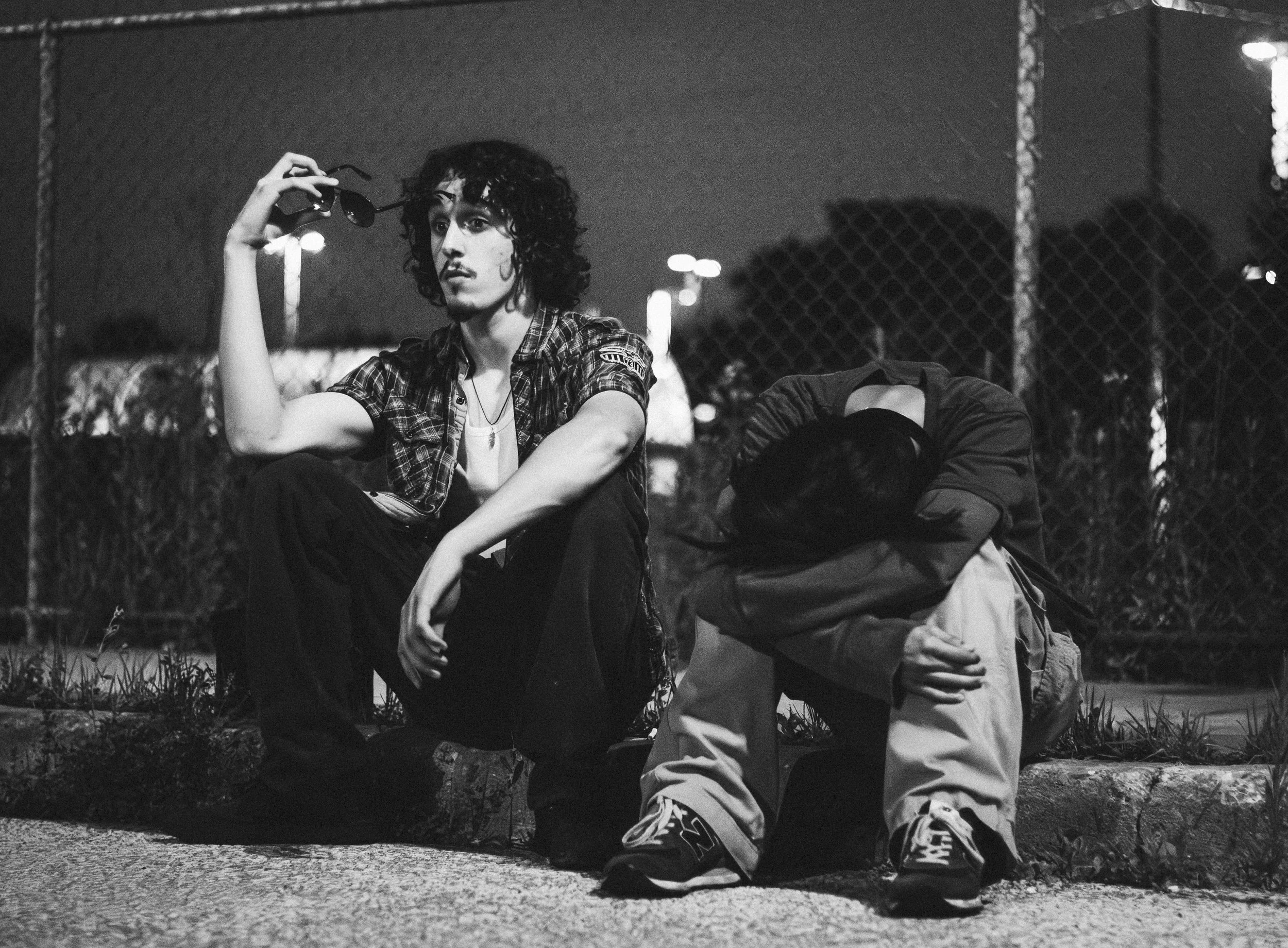 Black and white photo of two young women sitting on a curb at night. The woman on the left has curly hair, is holding sunglasses, and is looking thoughtfully into the distance. The woman on the right has dark hair, is hunched over with her arms wrapp