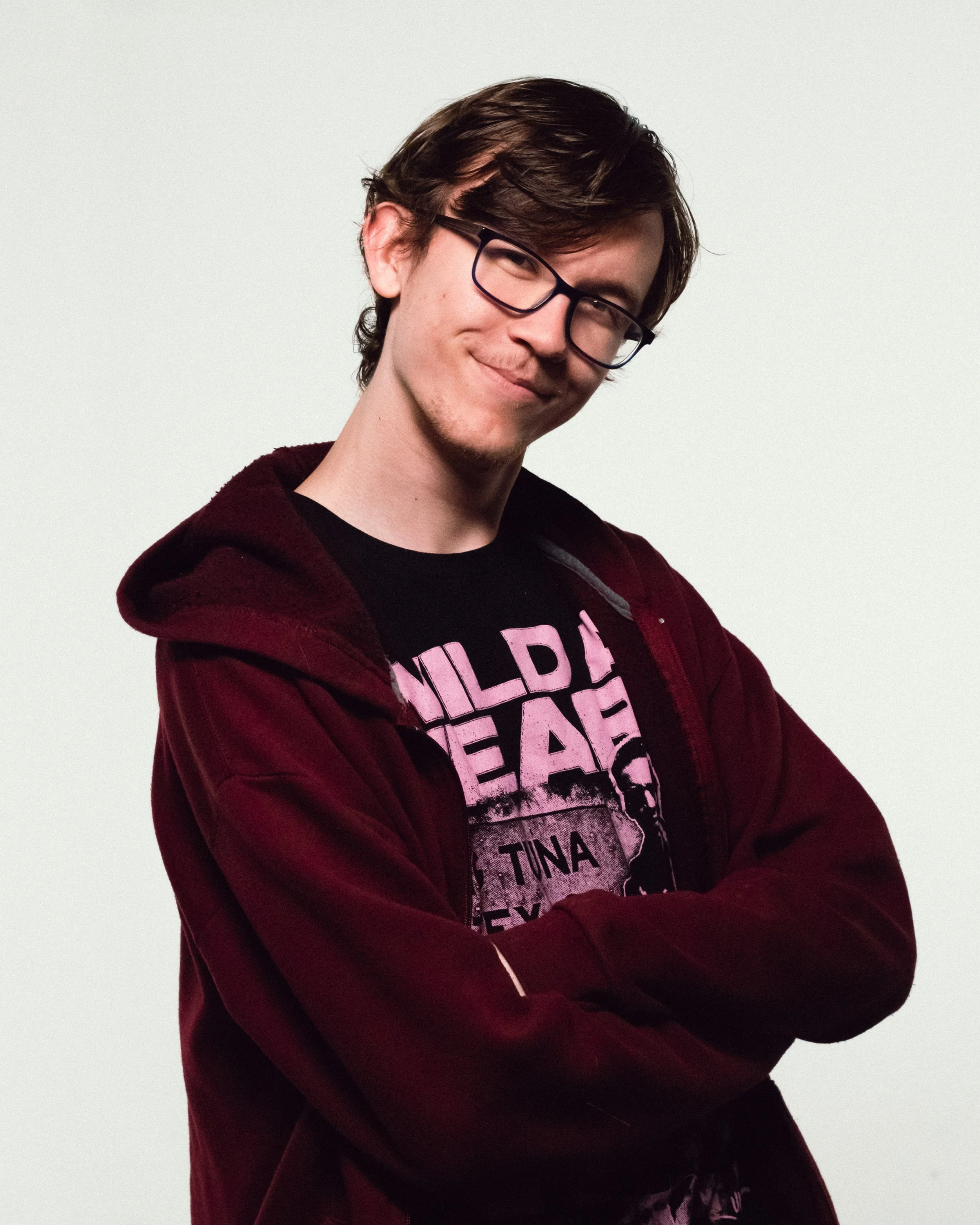 Young man with glasses smiling, wearing a black T-shirt with pink text and a maroon hoodie, arms crossed, against a plain background.