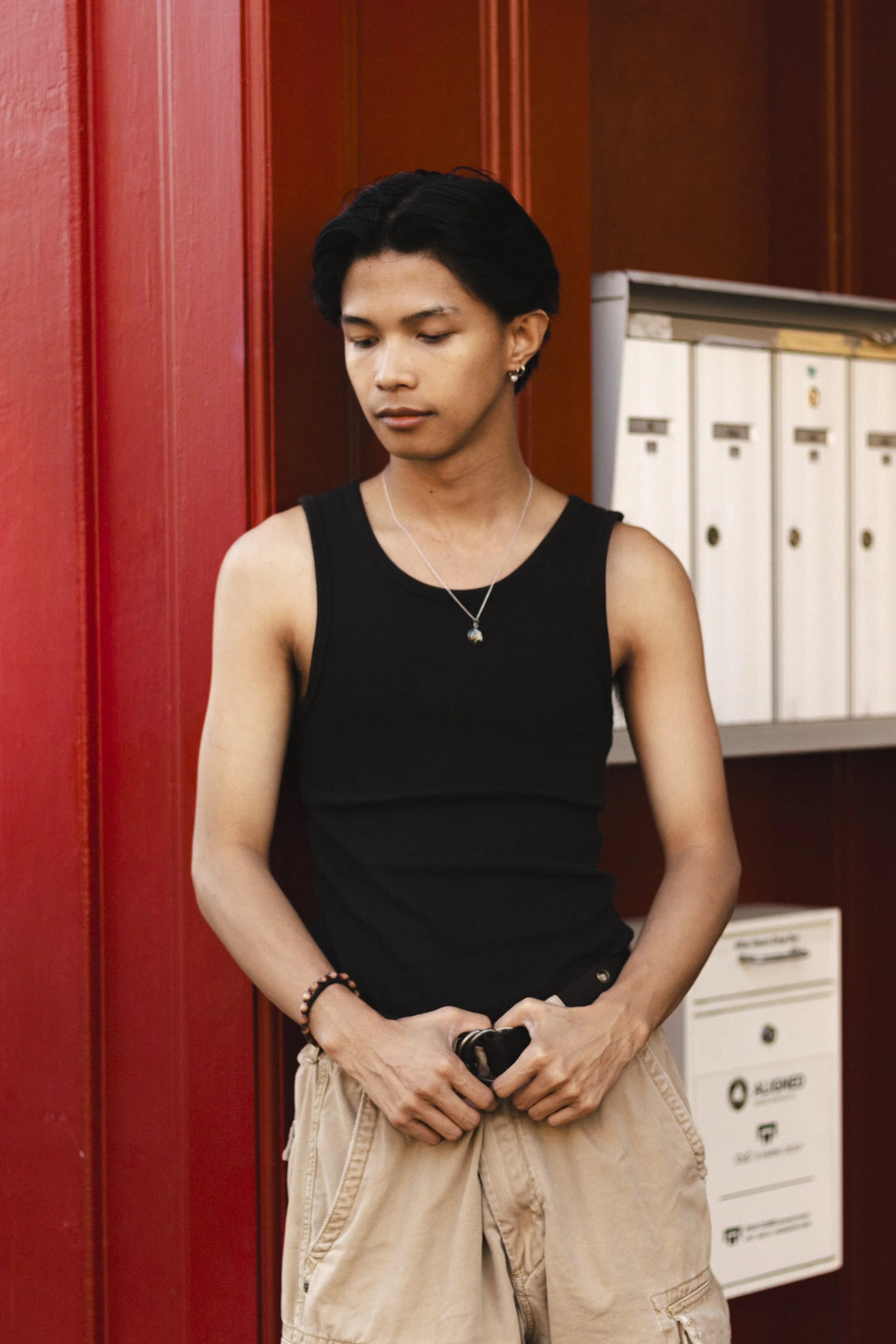 A young person with medium skin tone and black hair styled back, wearing a black sleeveless shirt, beige cargo shorts, a necklace, and a bracelet, standing against a red wall near mailboxes.