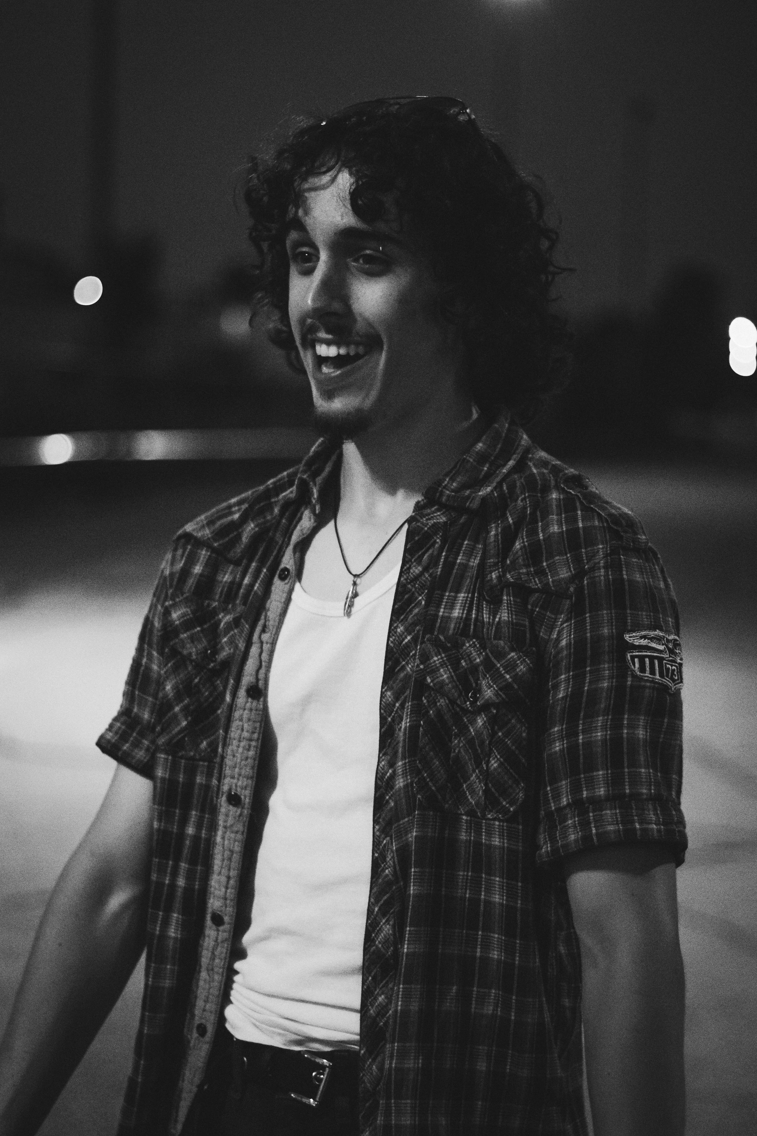 A young man with curly hair smiling and walking outdoors at night, wearing a plaid shirt over a white t-shirt, with a necklace and a patch on his sleeve.
