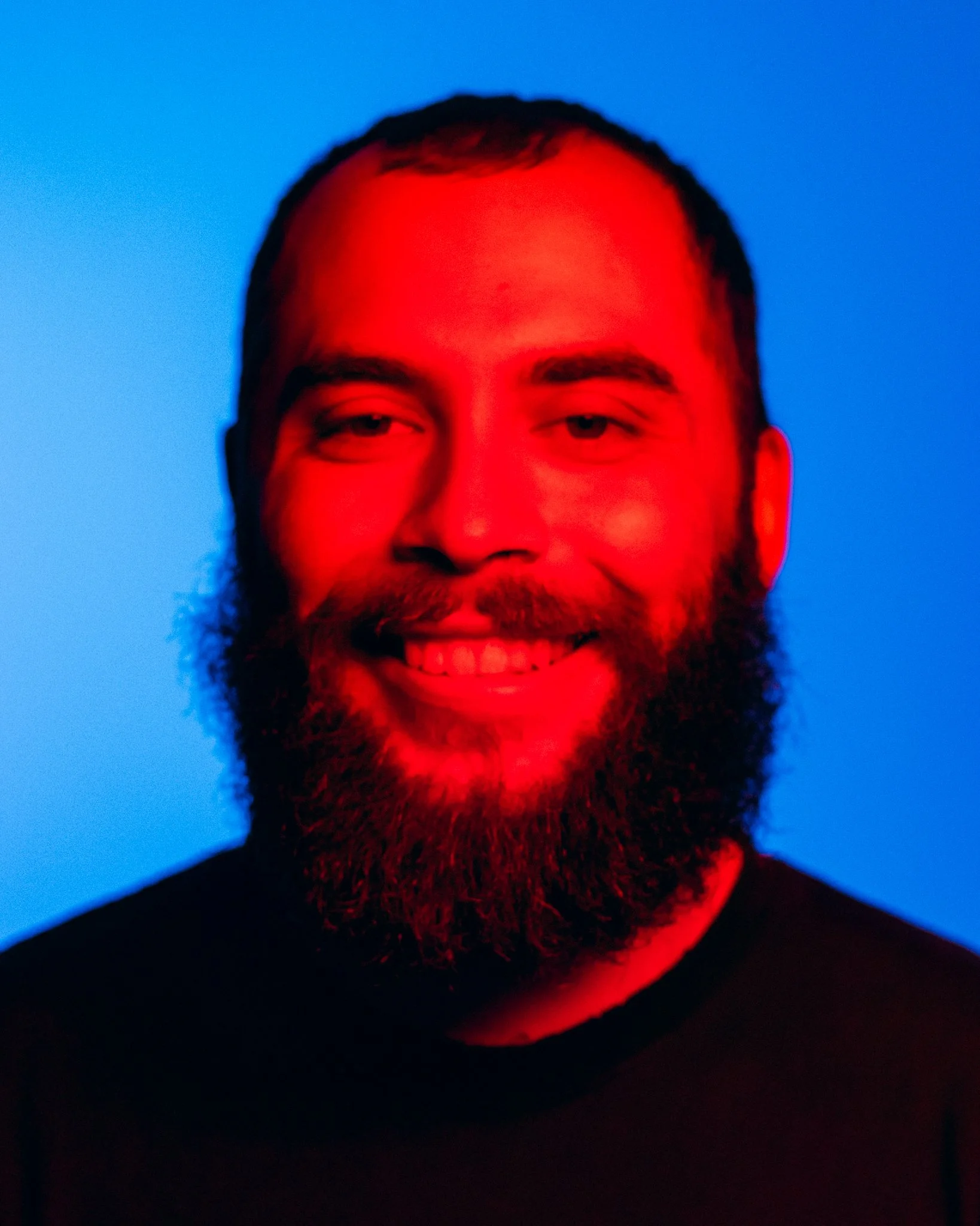 A man with a beard smiling against a blue background, illuminated by red and blue lighting.
