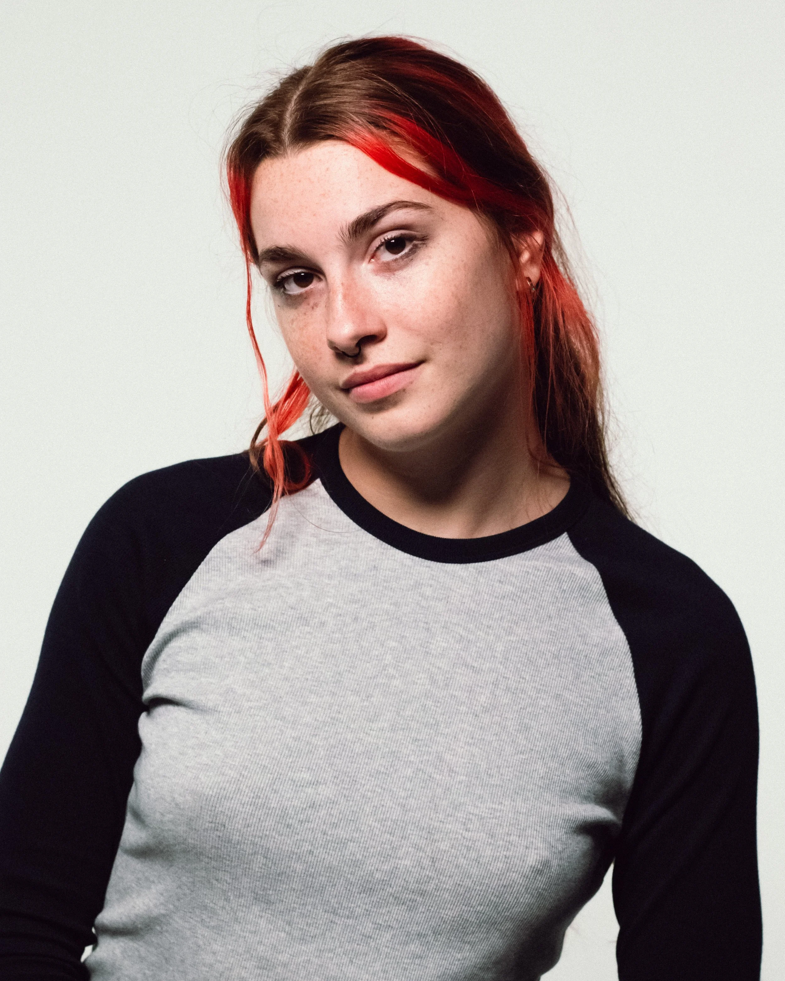A young woman with red hair and freckles wearing a black and gray baseball tee against a plain white background.