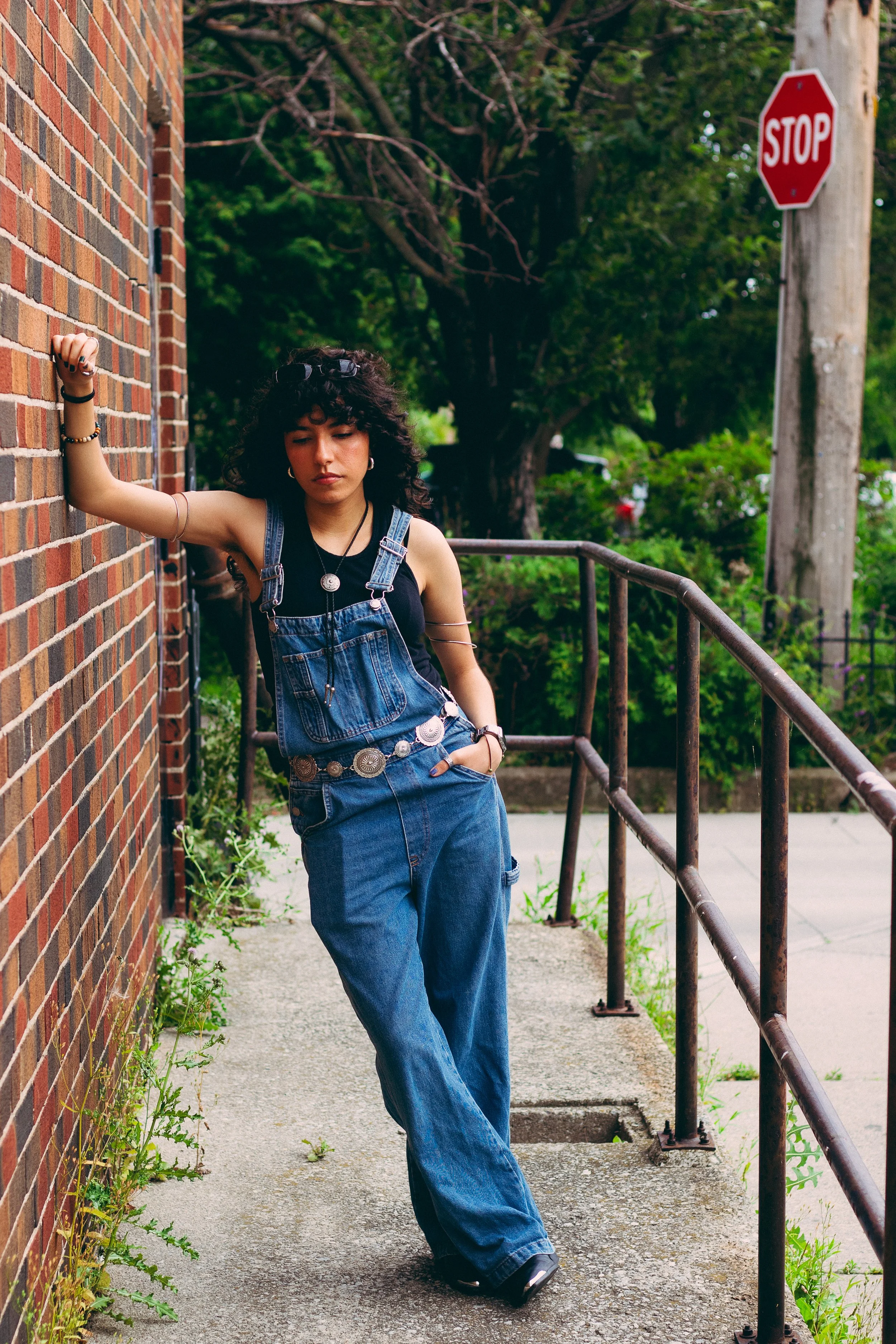 A young woman with curly dark hair wearing a black tank top, denim overalls, and jewelry, leaning against a brick wall on an outdoor sidewalk, with a stop sign and green trees in the background.