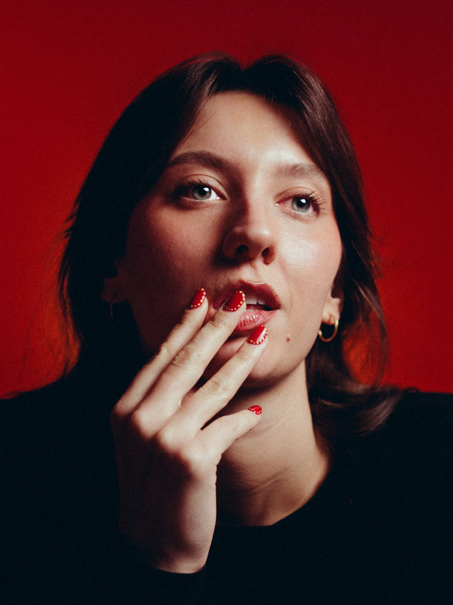 A woman with dark hair and blue eyes touching her lips with her fingers, painted with red nail polish, against a red background.