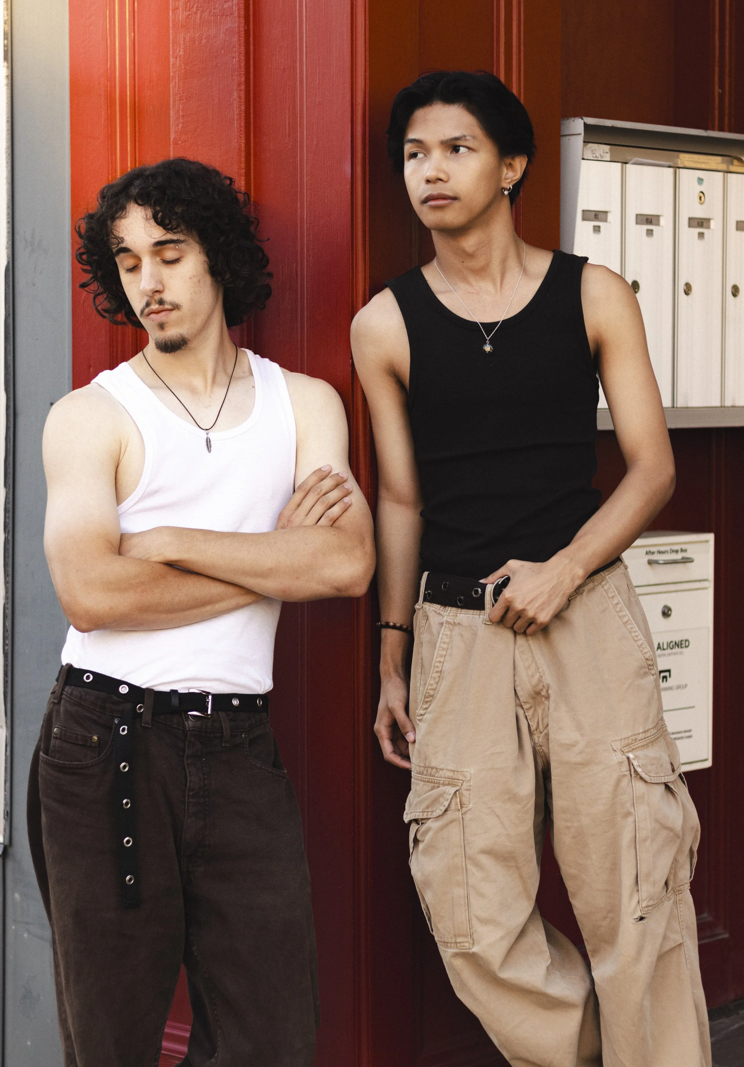 Two young men standing against a red wall with mailboxes, one with arms crossed and eyes closed, the other looking away with a serious expression.