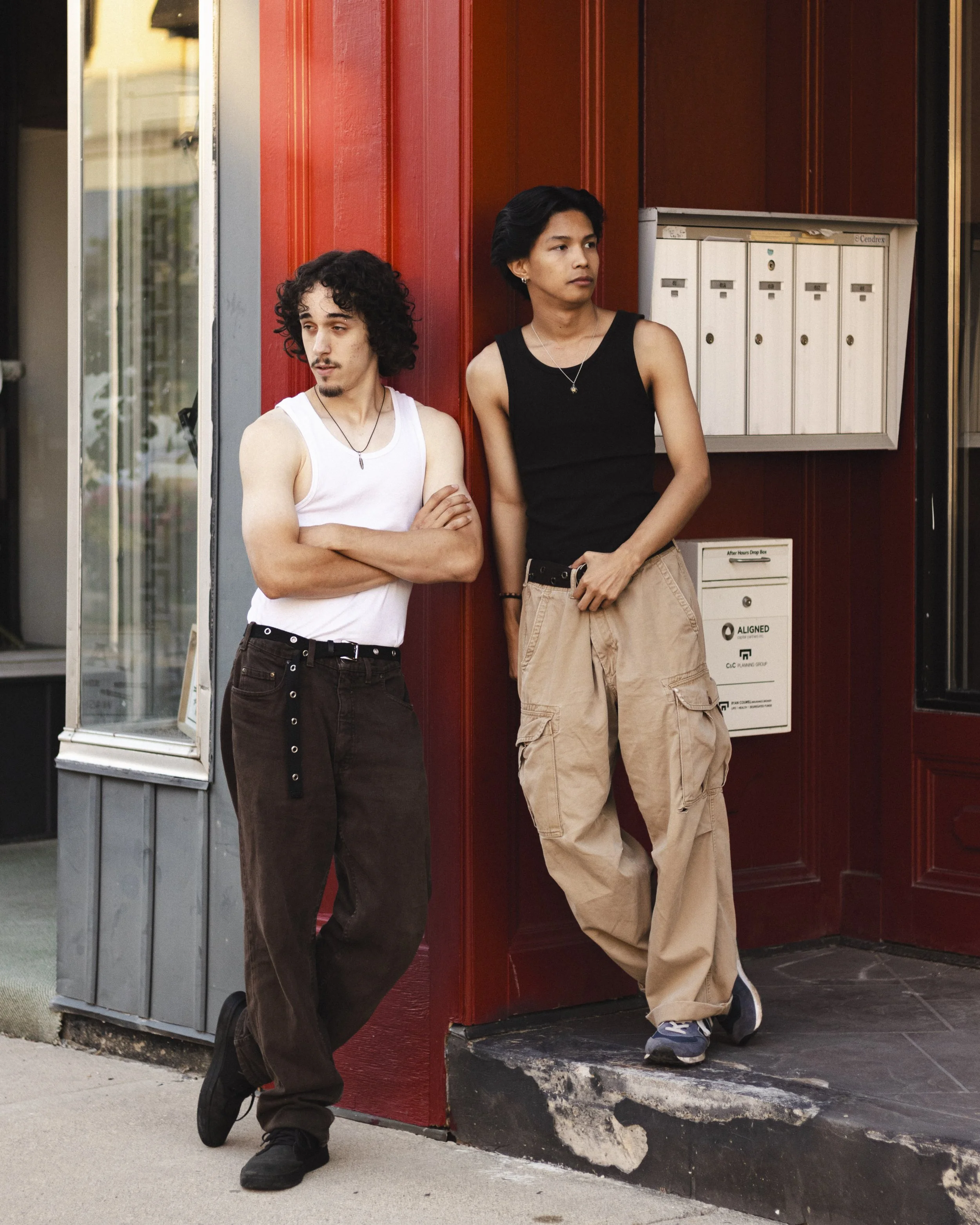 Two young people with tattoos and necklaces standing outside a red brick building near a row of mailboxes, one with arms crossed and the other with hands in pockets.