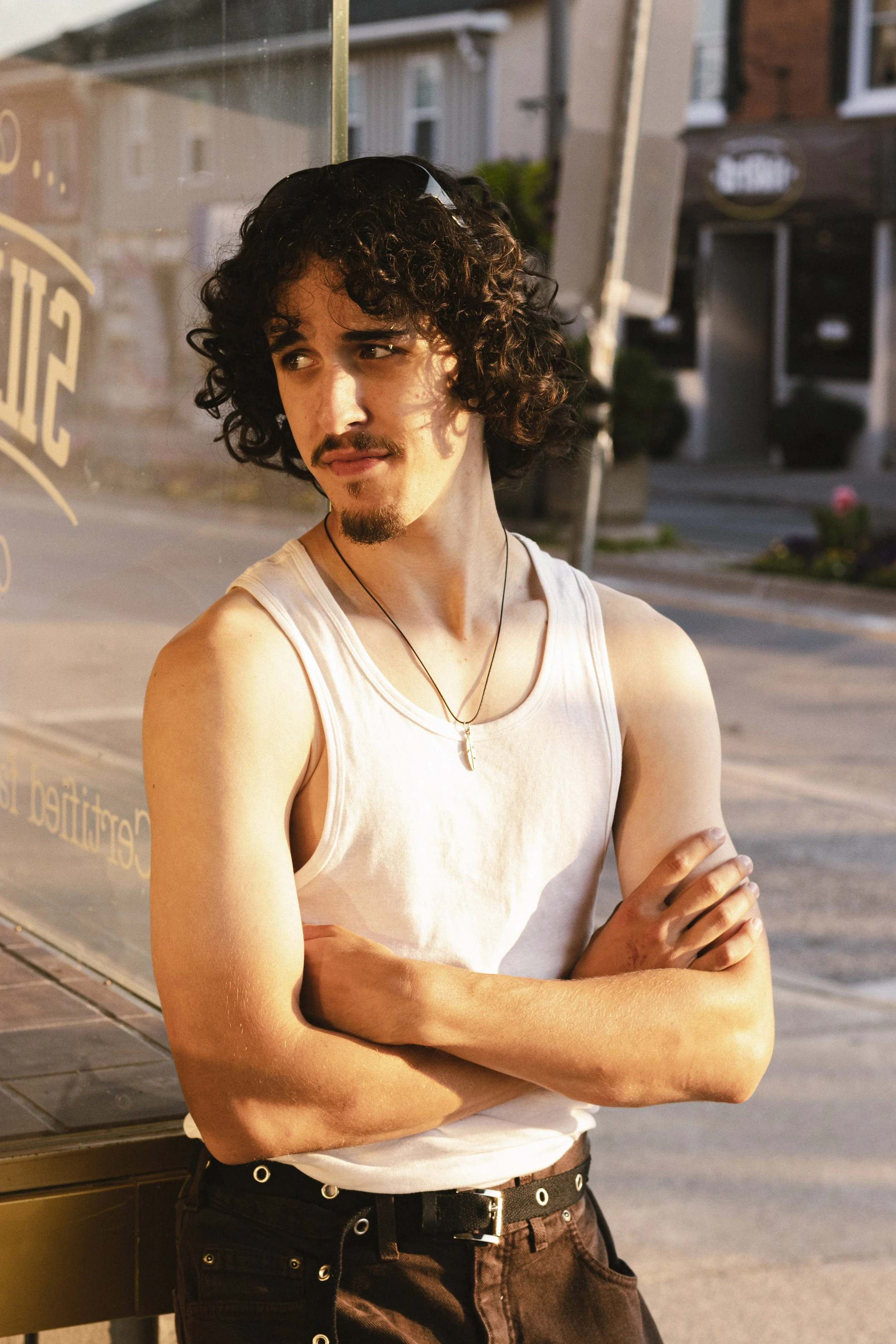 A young man with curly dark hair, mustache, and beard, wearing a sleeveless white shirt and necklace, standing with arms crossed outside during sunset or golden hour.