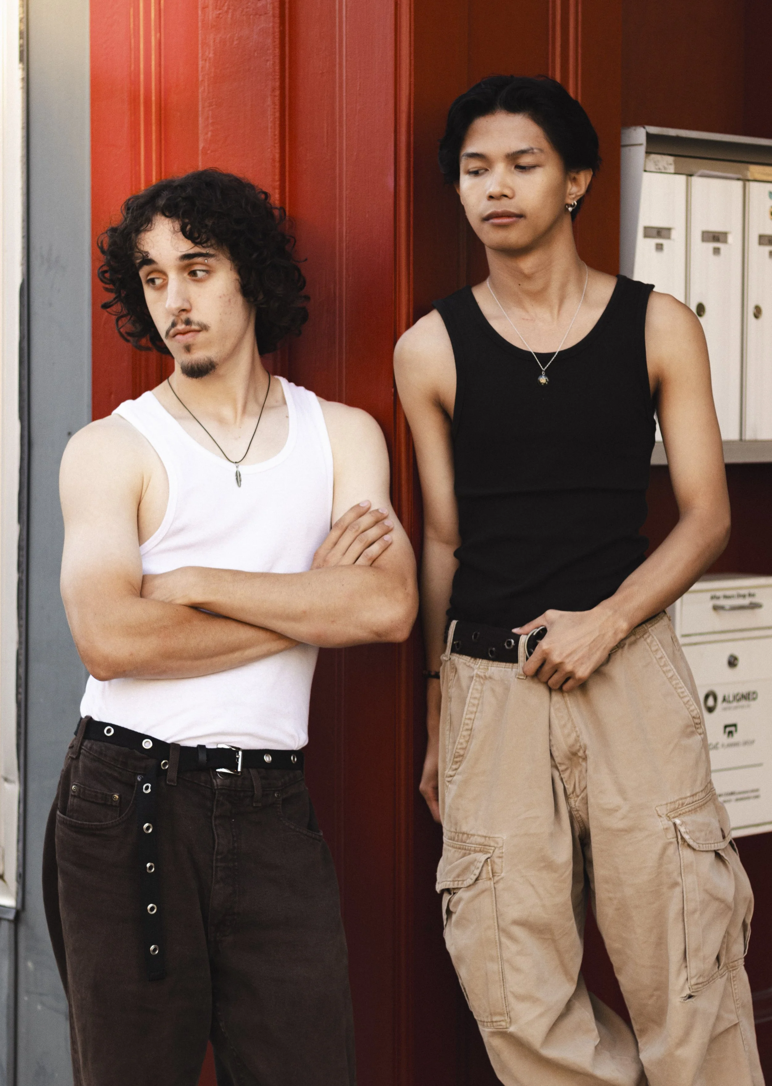 Two young men with slim builds standing against a red panel, one with muscular arms, wearing a white tank top and black pants, arms crossed, the other with a longer stance, wearing a black tank top and beige cargo pants, with short dark hair, both lo