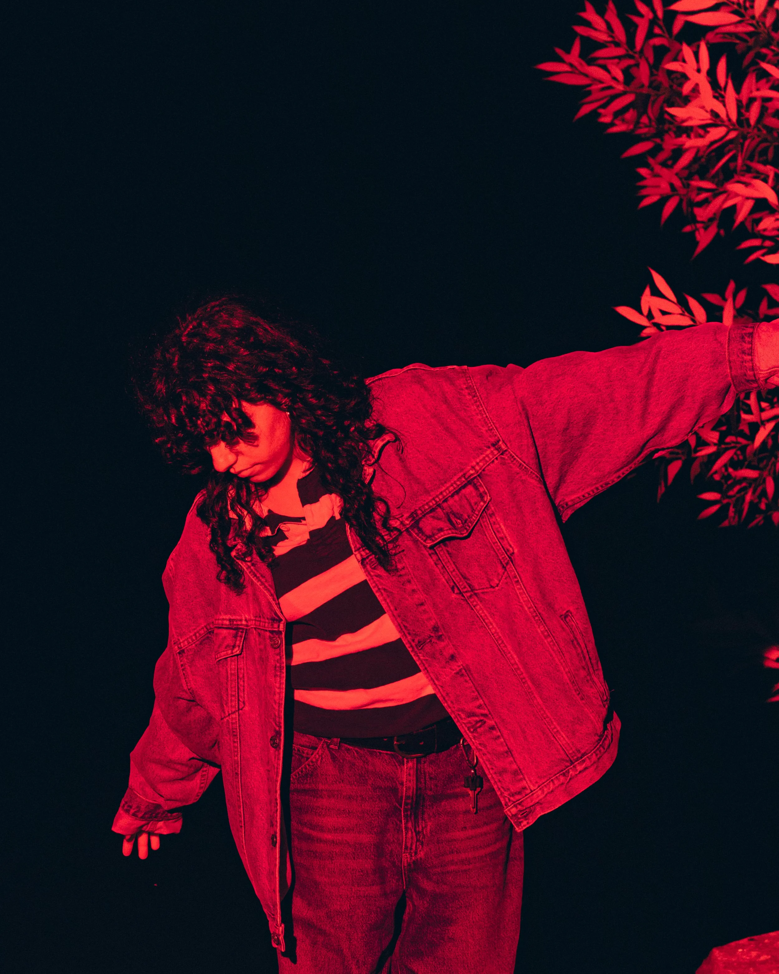 Person with curly hair wearing a striped shirt and denim jacket, standing outdoors at night with red lighting and a tree with red leaves in the background.