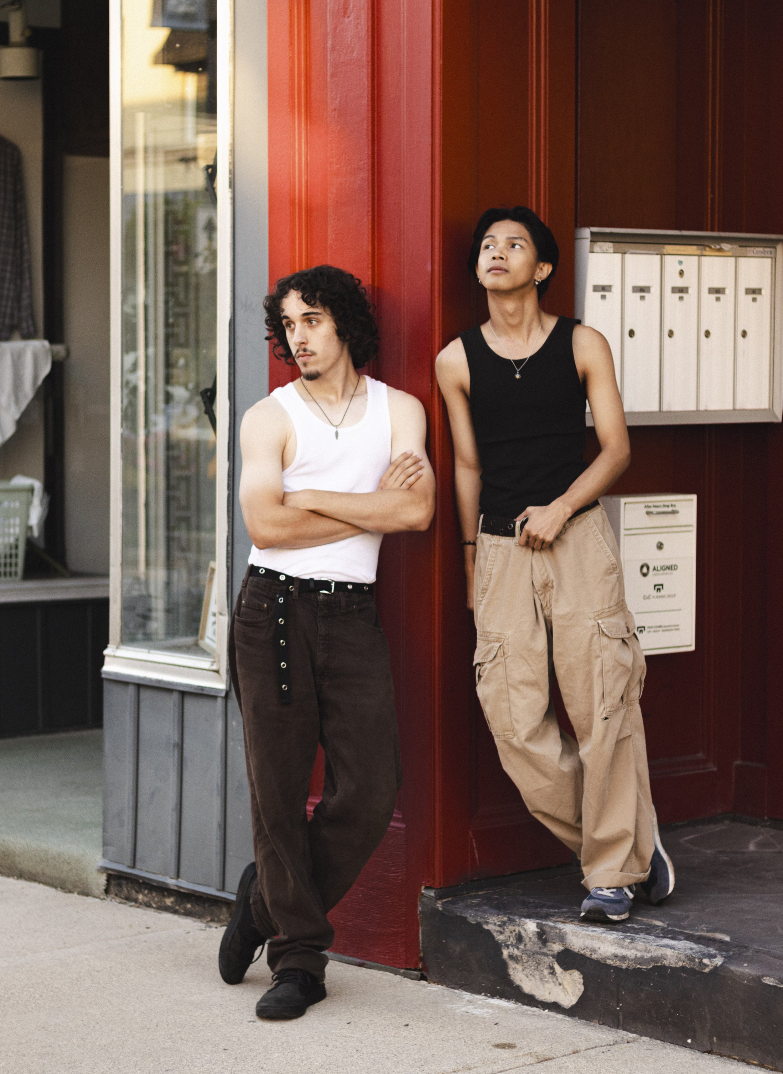 Two young men with arm tattoos leaning against a building corner outside, both with long hair, one in a white tank top and the other in a black tank top, with a background of red wood paneling and mailboxes.