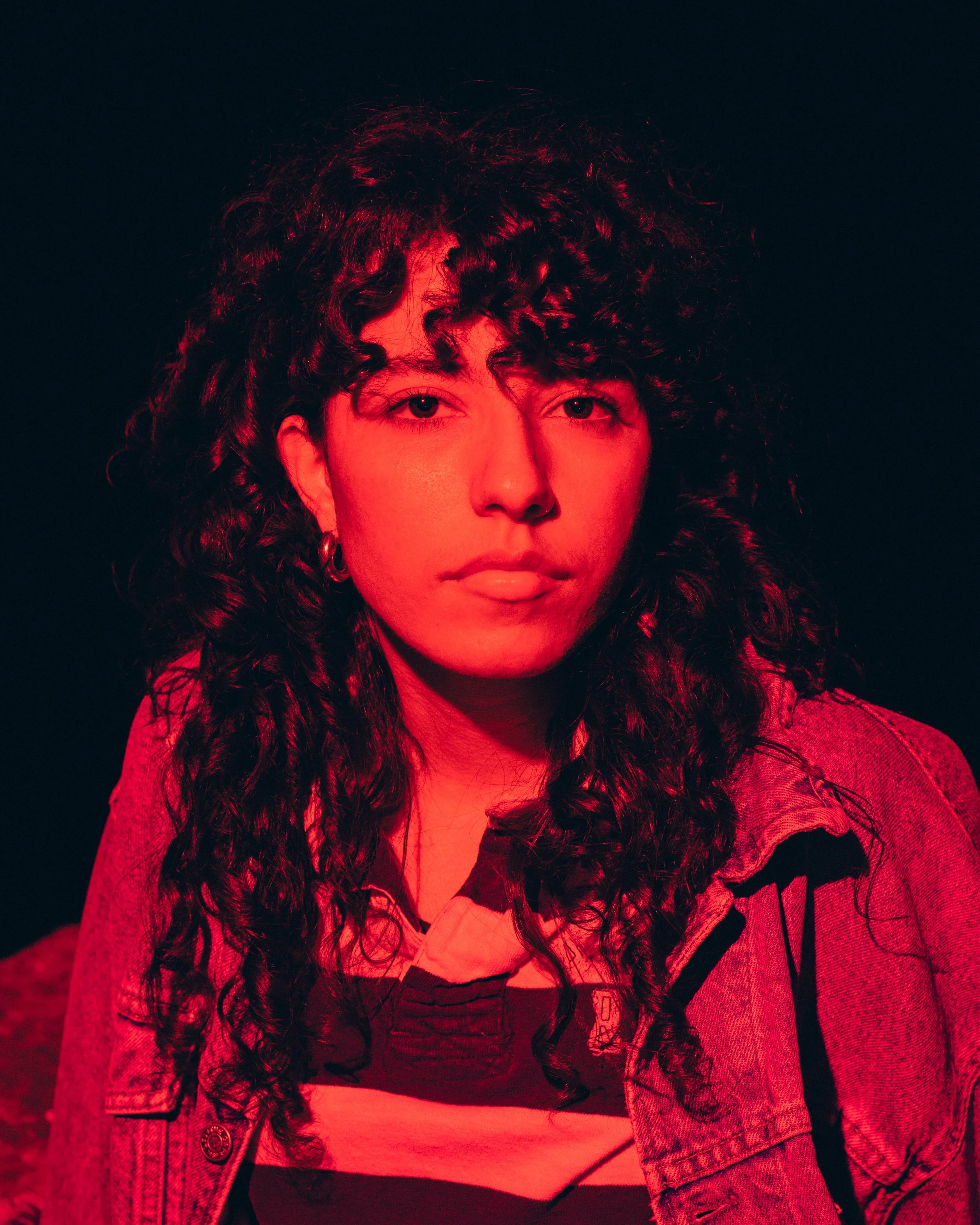 A woman with curly hair wearing a striped shirt and denim jacket, lit with red lighting against a dark background.