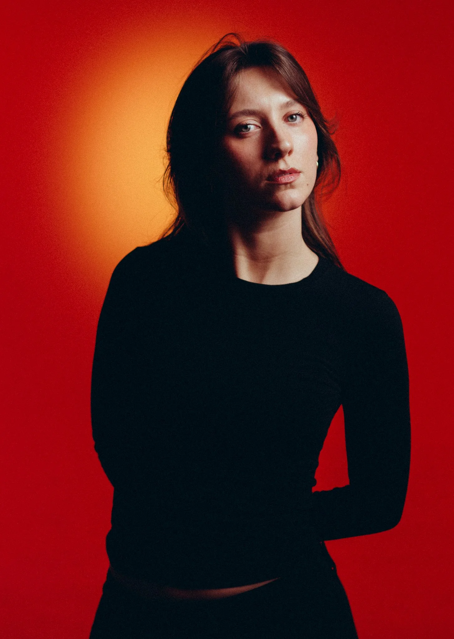 A portrait of a woman with light skin and brown hair, wearing a black top, standing against a red and orange gradient background.