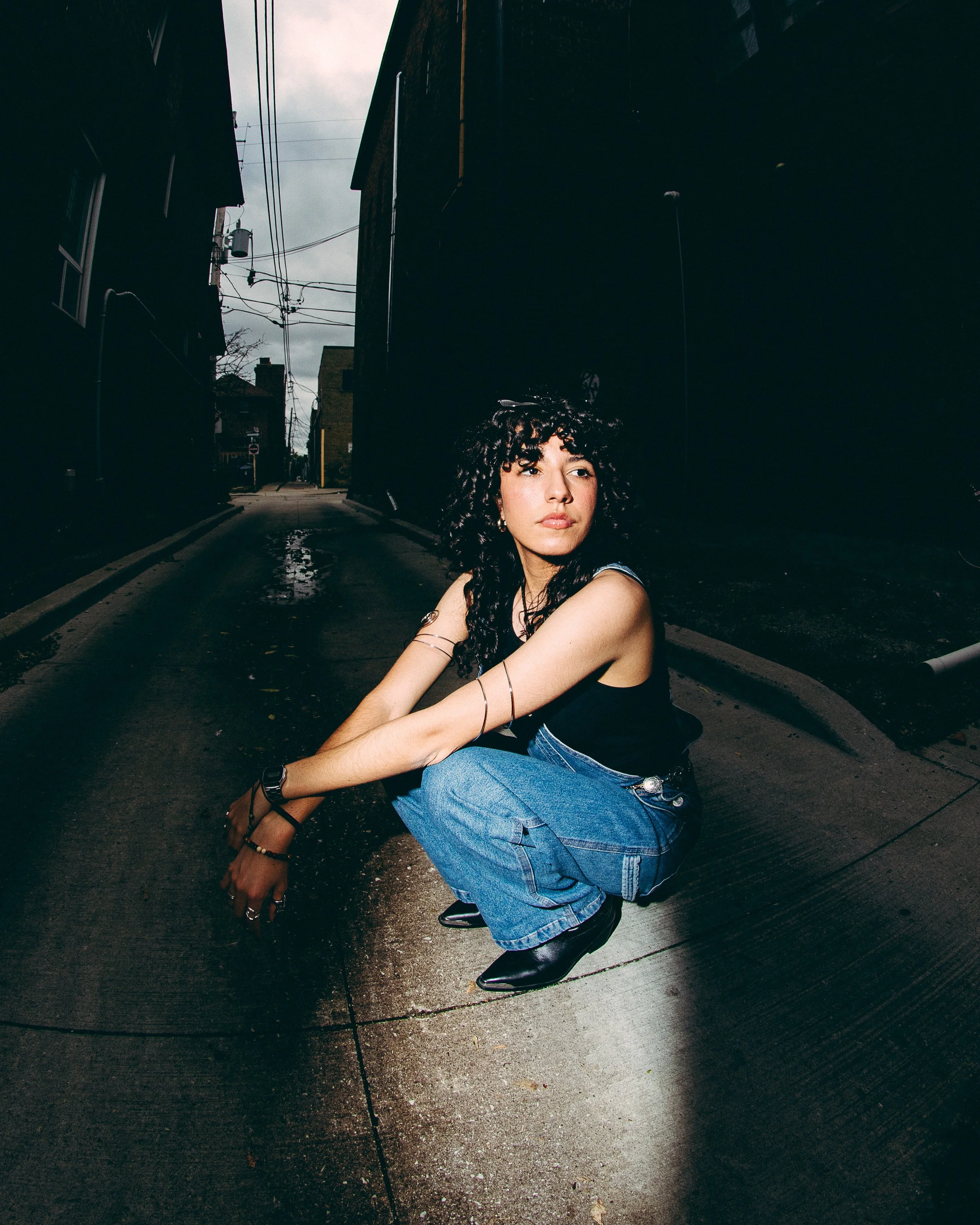A woman with curly black hair squatting on a city street in a narrow alley, wearing a black tank top, blue jeans, and black boots, with dangling earrings and various bracelets.