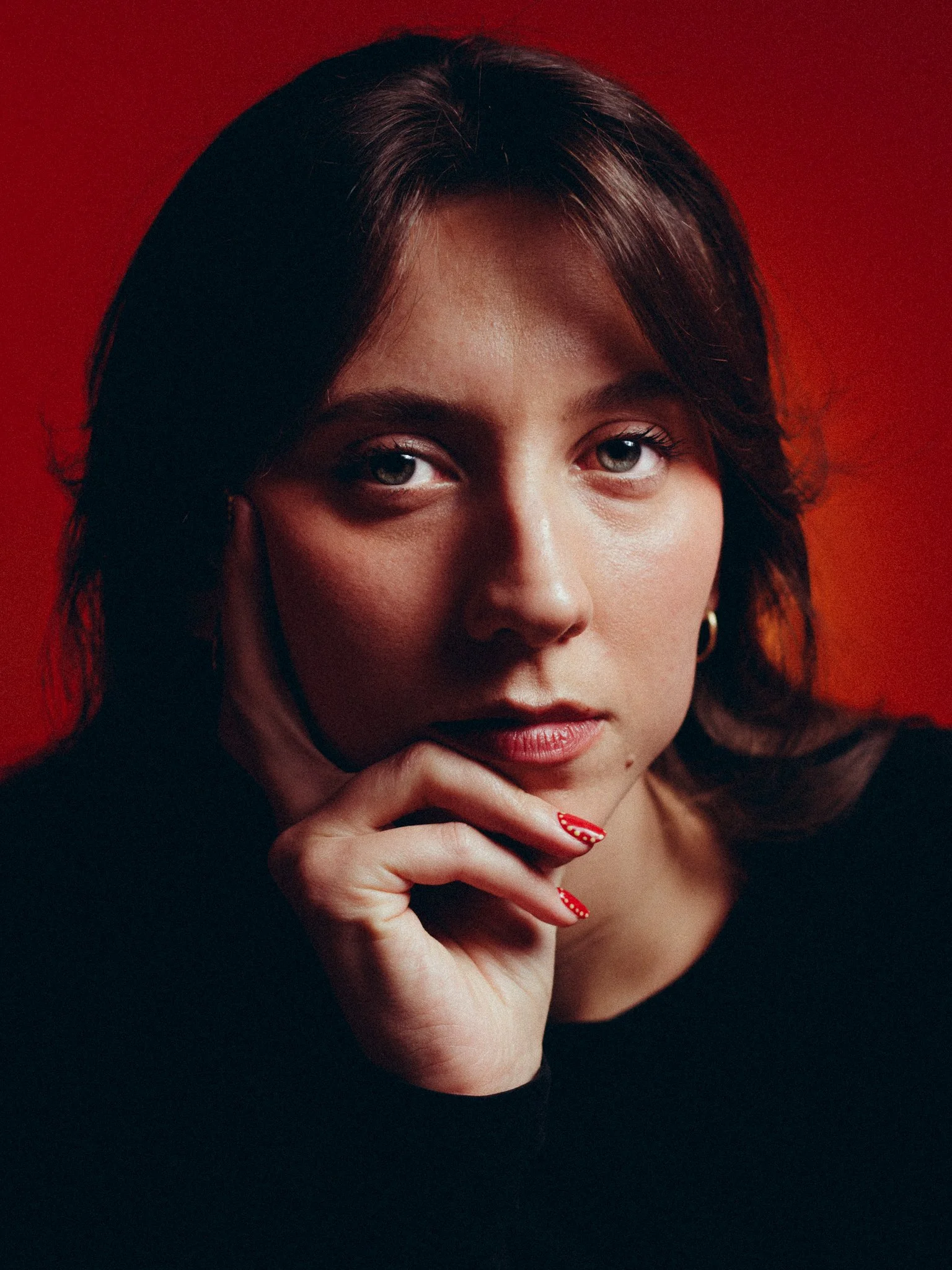 Close-up portrait of a young woman with dark hair, wearing minimal makeup, a black top, and red nail polish, resting her chin on her hand against a red background.