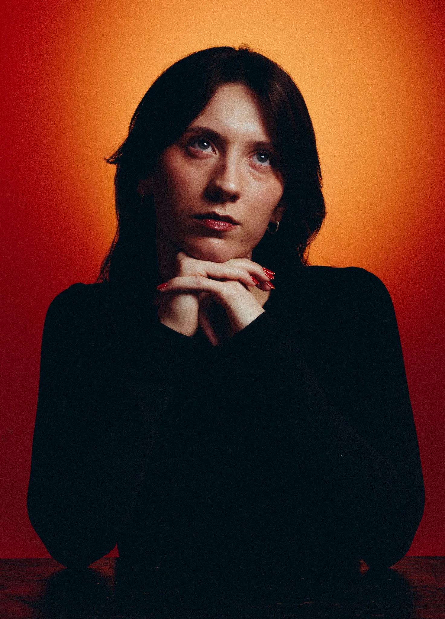 A woman with dark hair and red nail polish, resting her chin on her hands, looking slightly upwards. She is wearing a black top, with an orange gradient background.