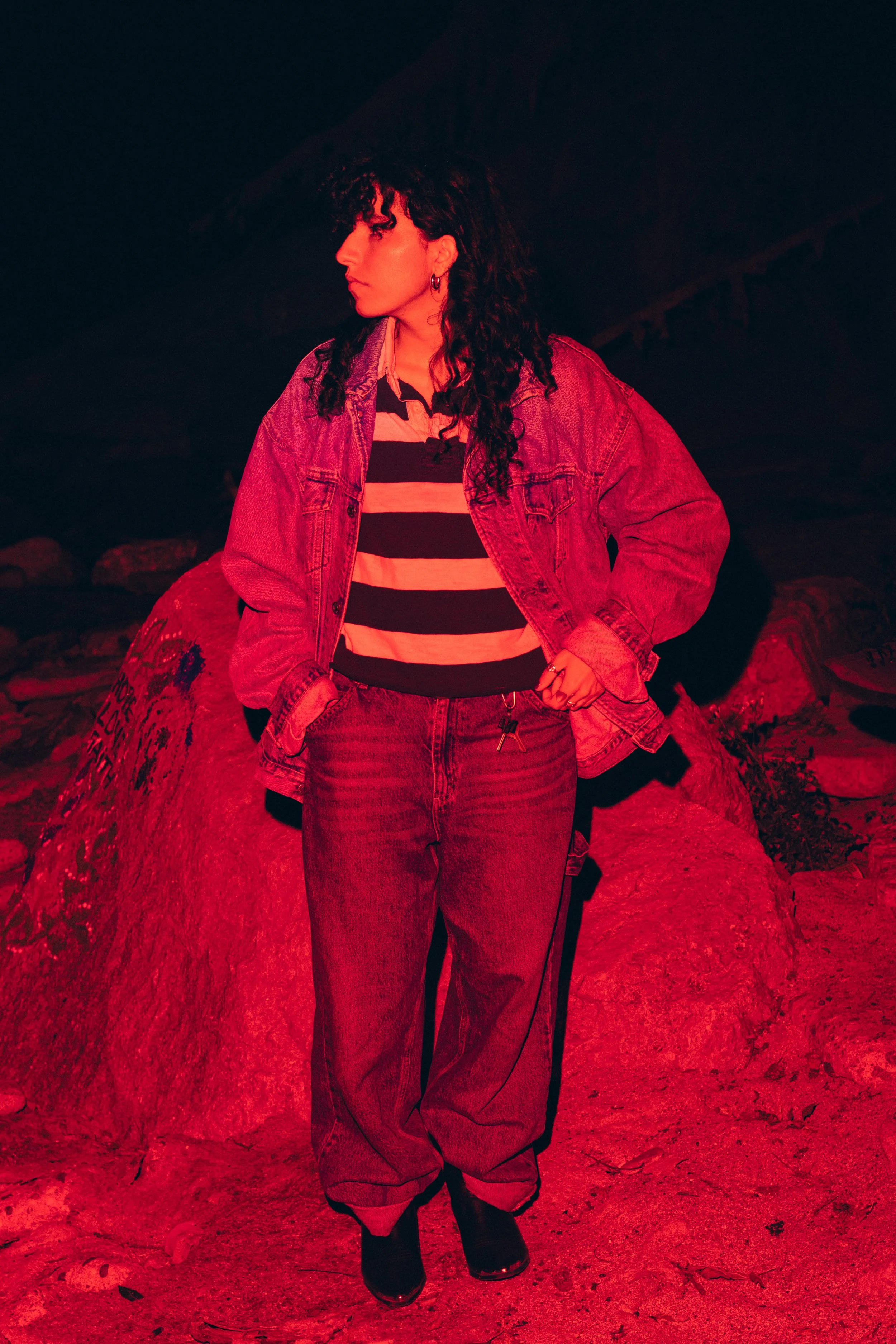 A woman with curly hair wearing a denim jacket, striped shirt, and jeans standing outdoors on rocky terrain at night with a dark background.