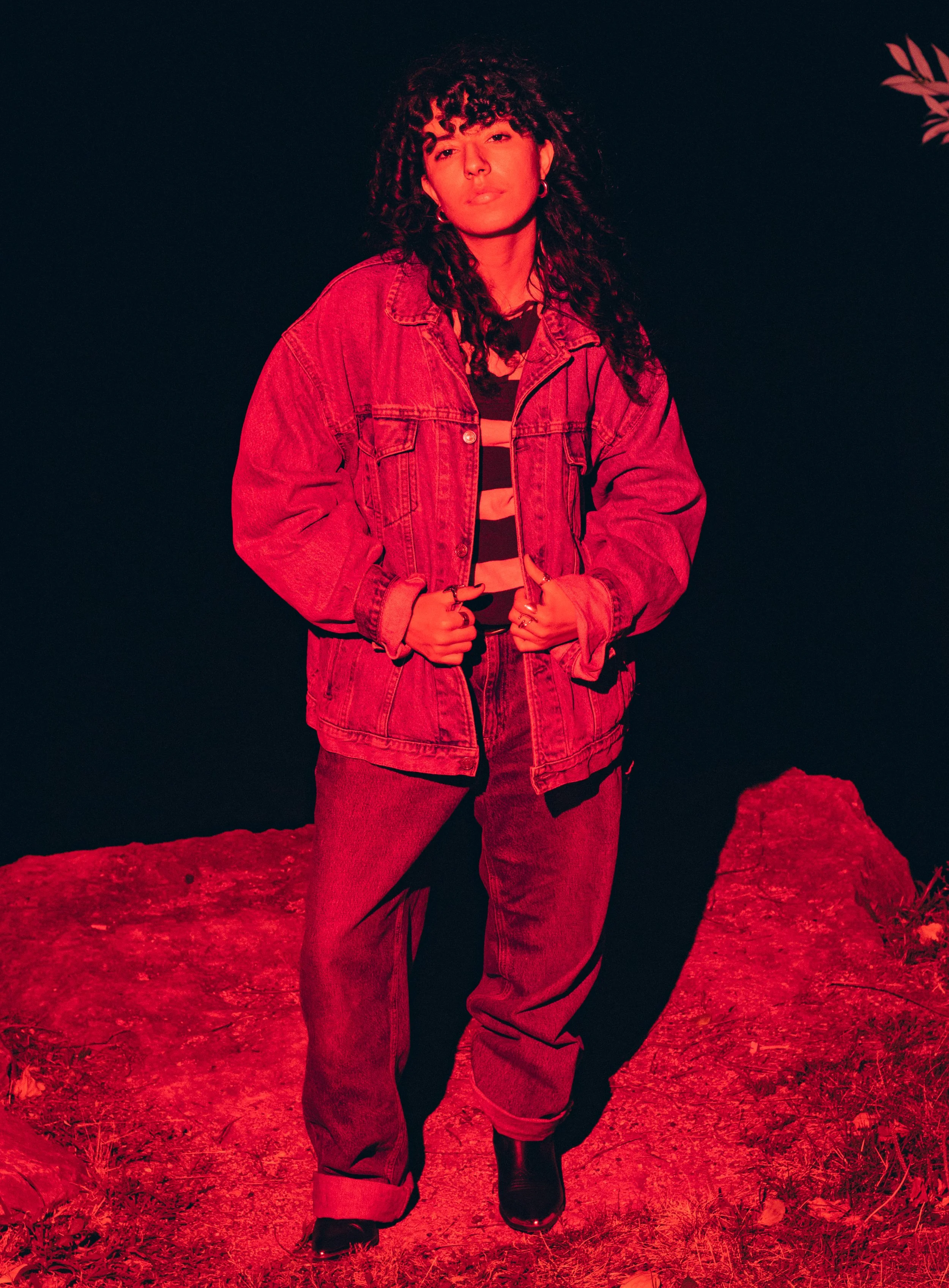 A woman with curly hair wearing a denim jacket, striped shirt, and jeans, standing outdoors at night on a rocky ground, illuminated with red lighting.