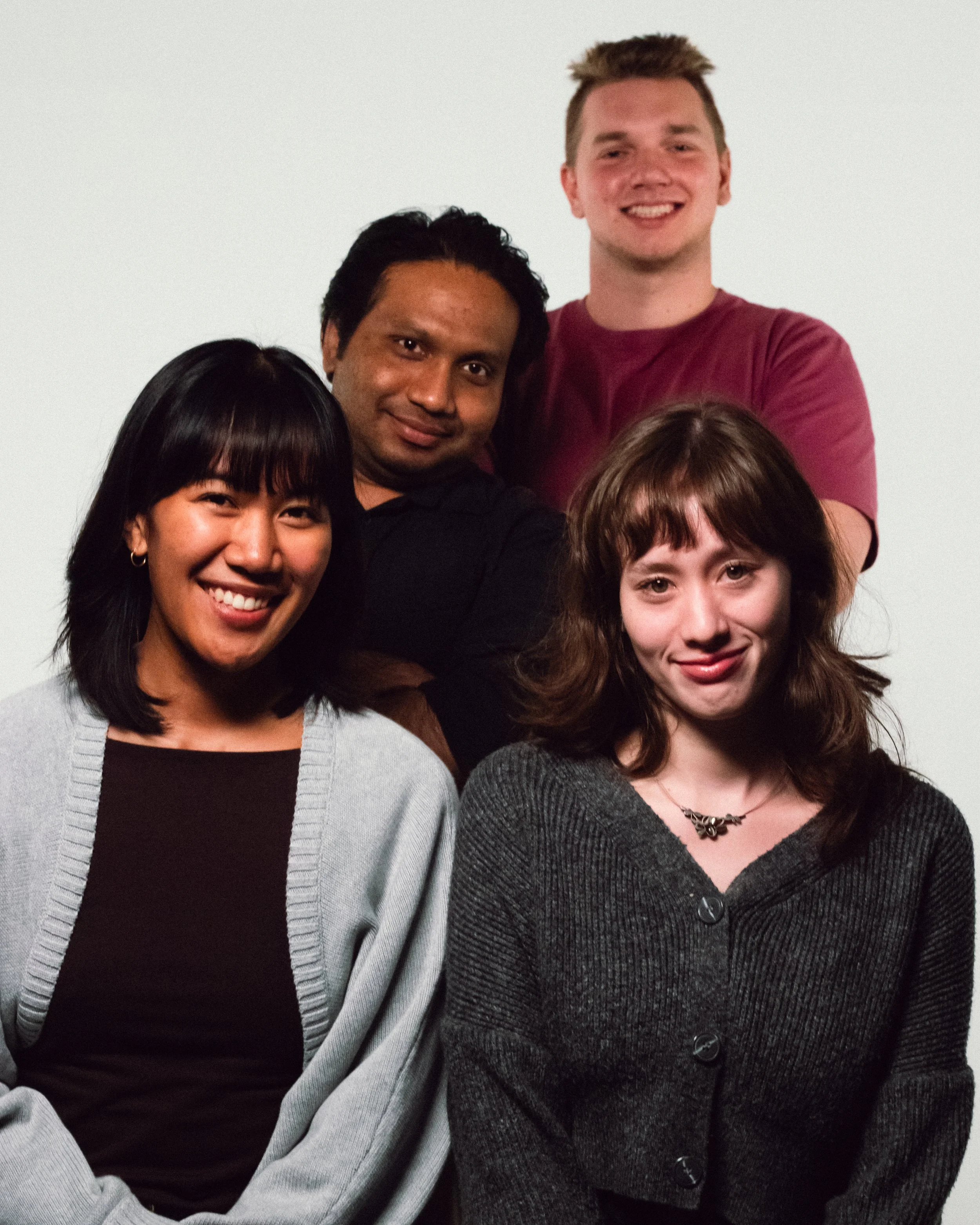 A diverse group of five young adults smiling, posing against a plain light-colored background. Two women are seated in the front, and two men and one woman are standing behind them.