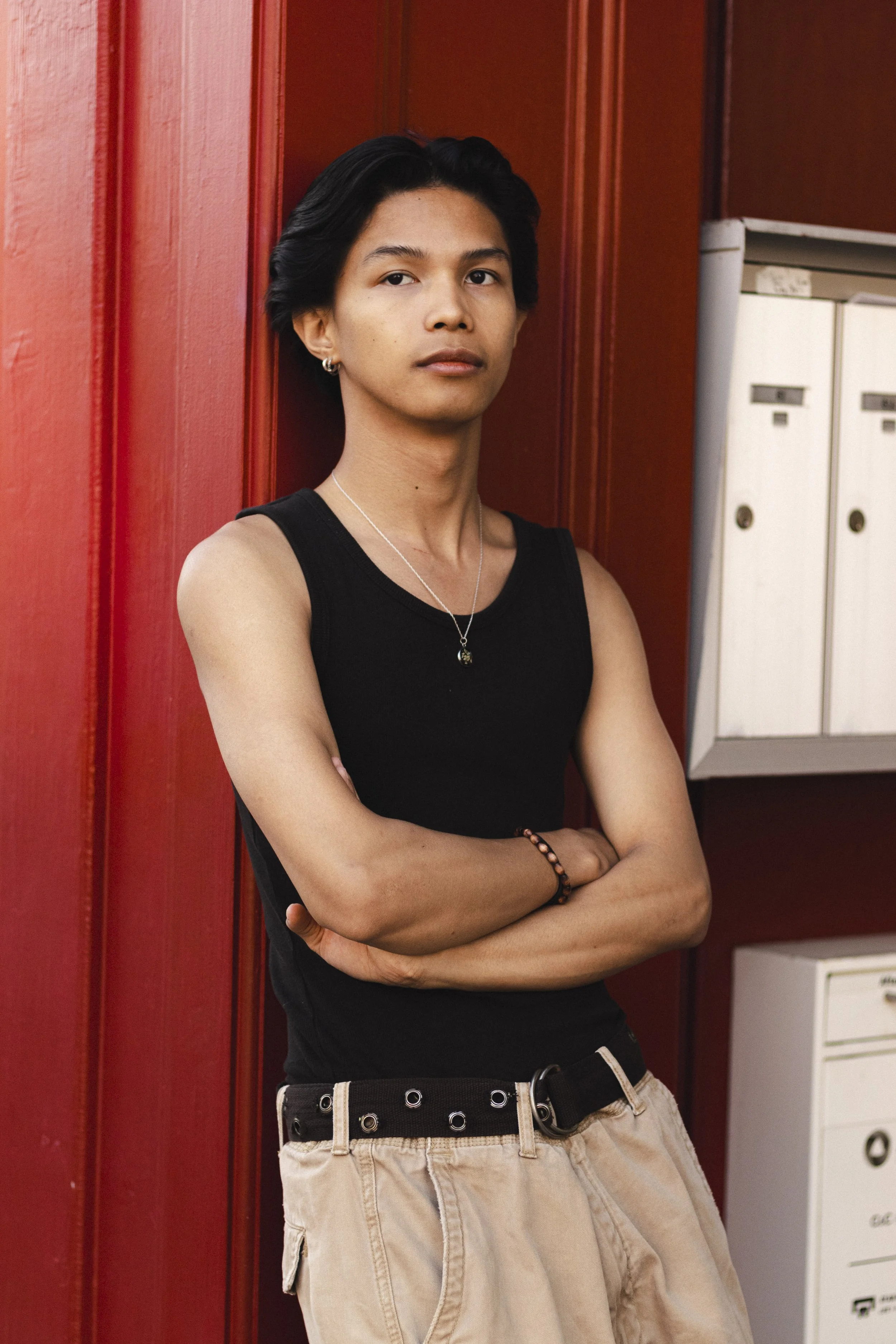 A young person with dark hair, wearing a black tank top, standing with arms crossed, in front of a red wall with mailboxes.