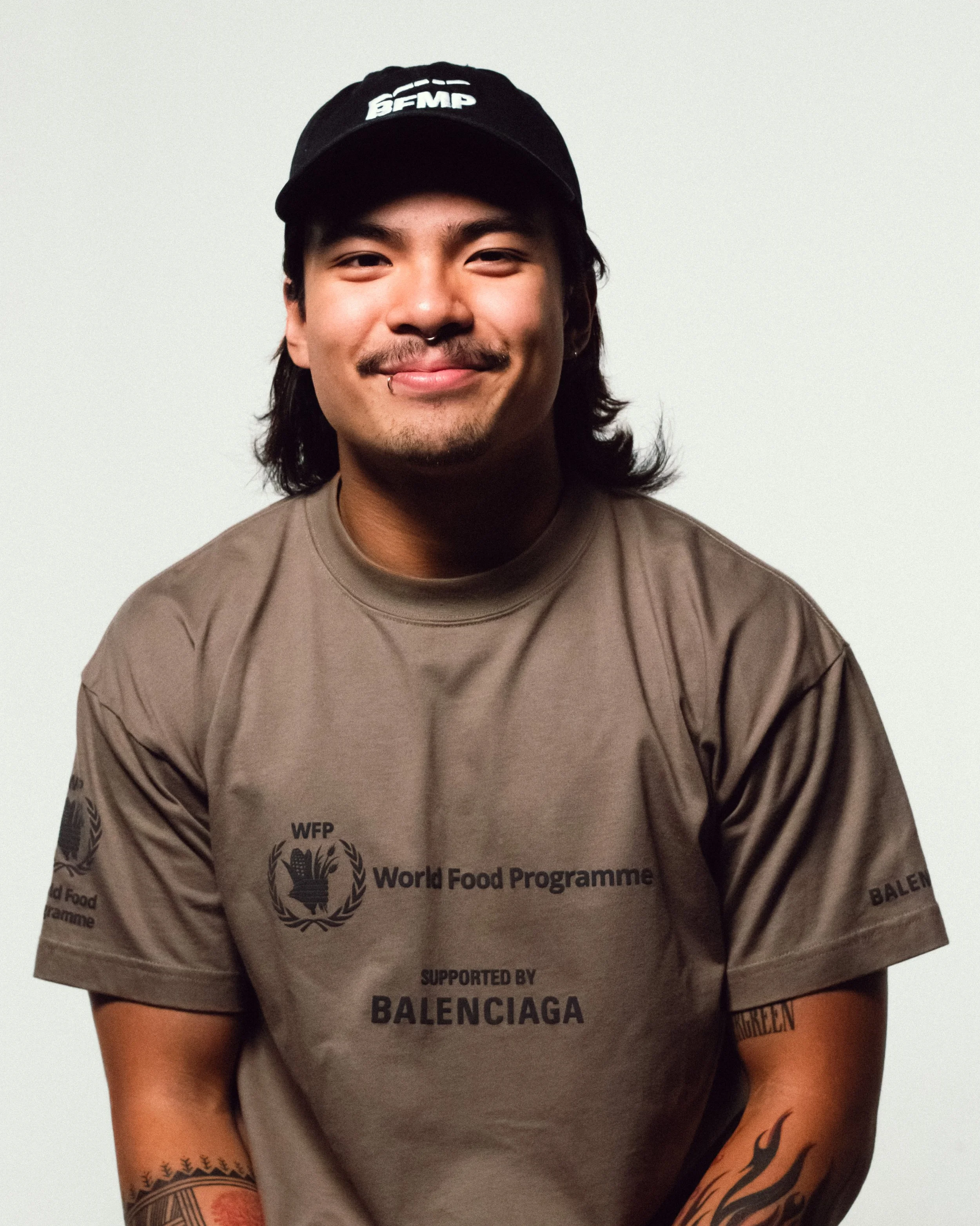 A young man with a mustache and goatee wearing a black cap and a brown T-shirt with 'World Food Programme' and 'Supported by Balenciaga' printed on it. He has tattoos on his arms and is smiling at the camera.