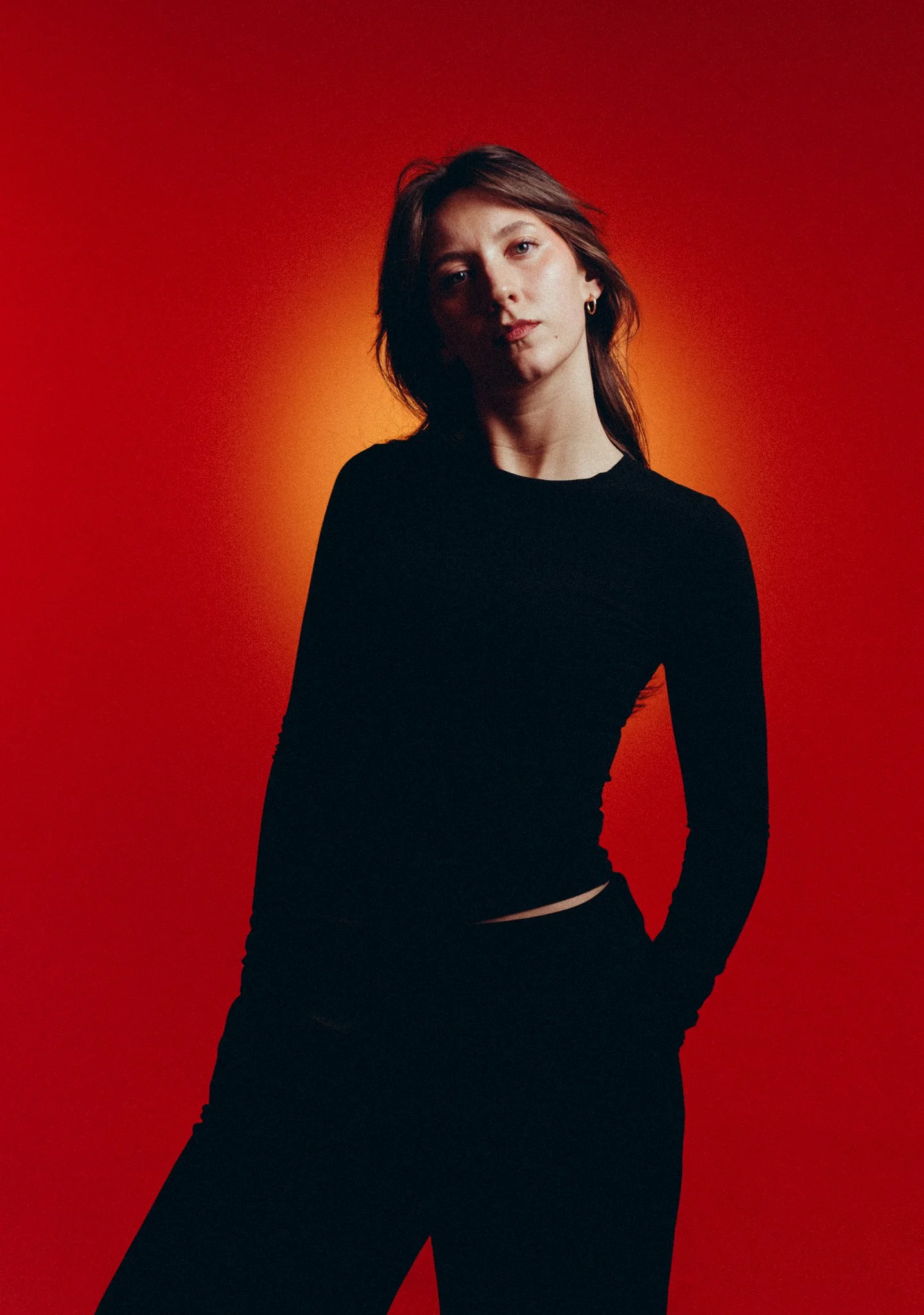 A woman with brown hair wearing a black long-sleeve top and black pants poses against an orange background, with her left hand in her pocket.