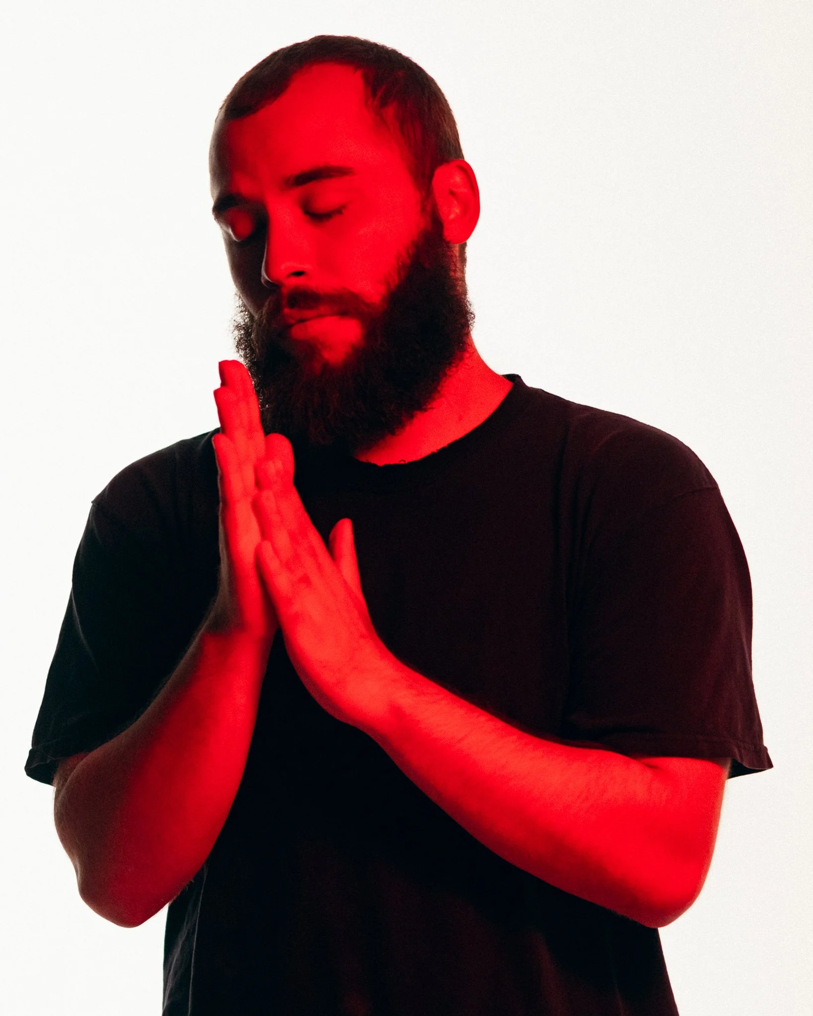 A man with a beard and closed eyes standing with hands pressed together in a prayer position, illuminated by red light.