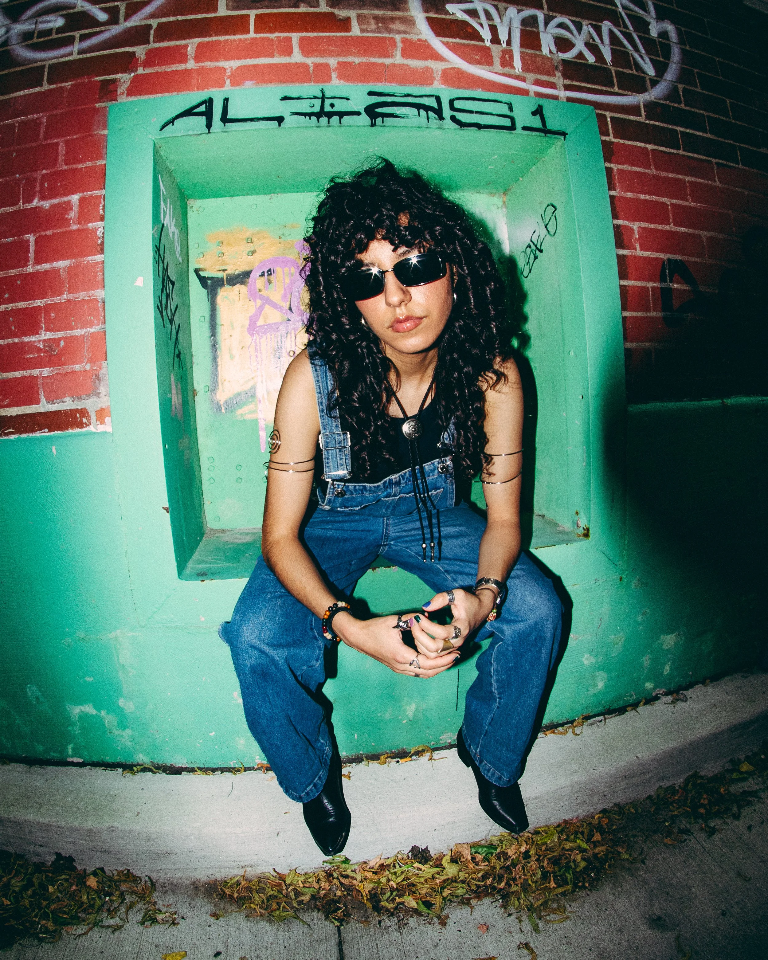 A woman with curly black hair, wearing black sunglasses, a black sleeveless top, blue denim overalls, and multiple rings and bracelets, sits on a green concrete ledge in an urban setting with graffiti on the brick wall behind her.