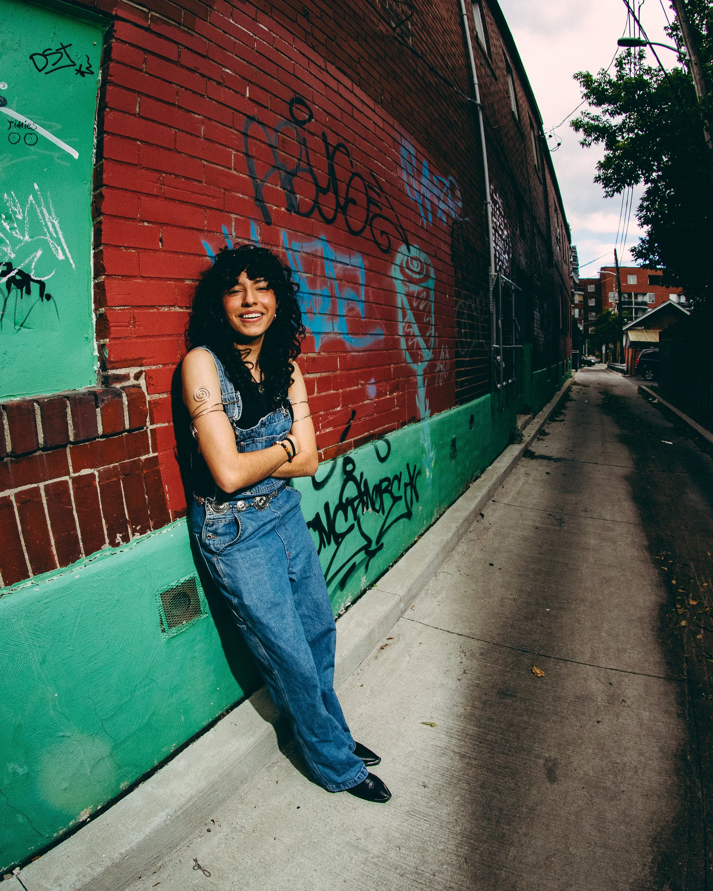 Young woman with curly black hair smiling and standing with arms crossed on a sidewalk next to a graffiti-covered brick wall in an urban setting.