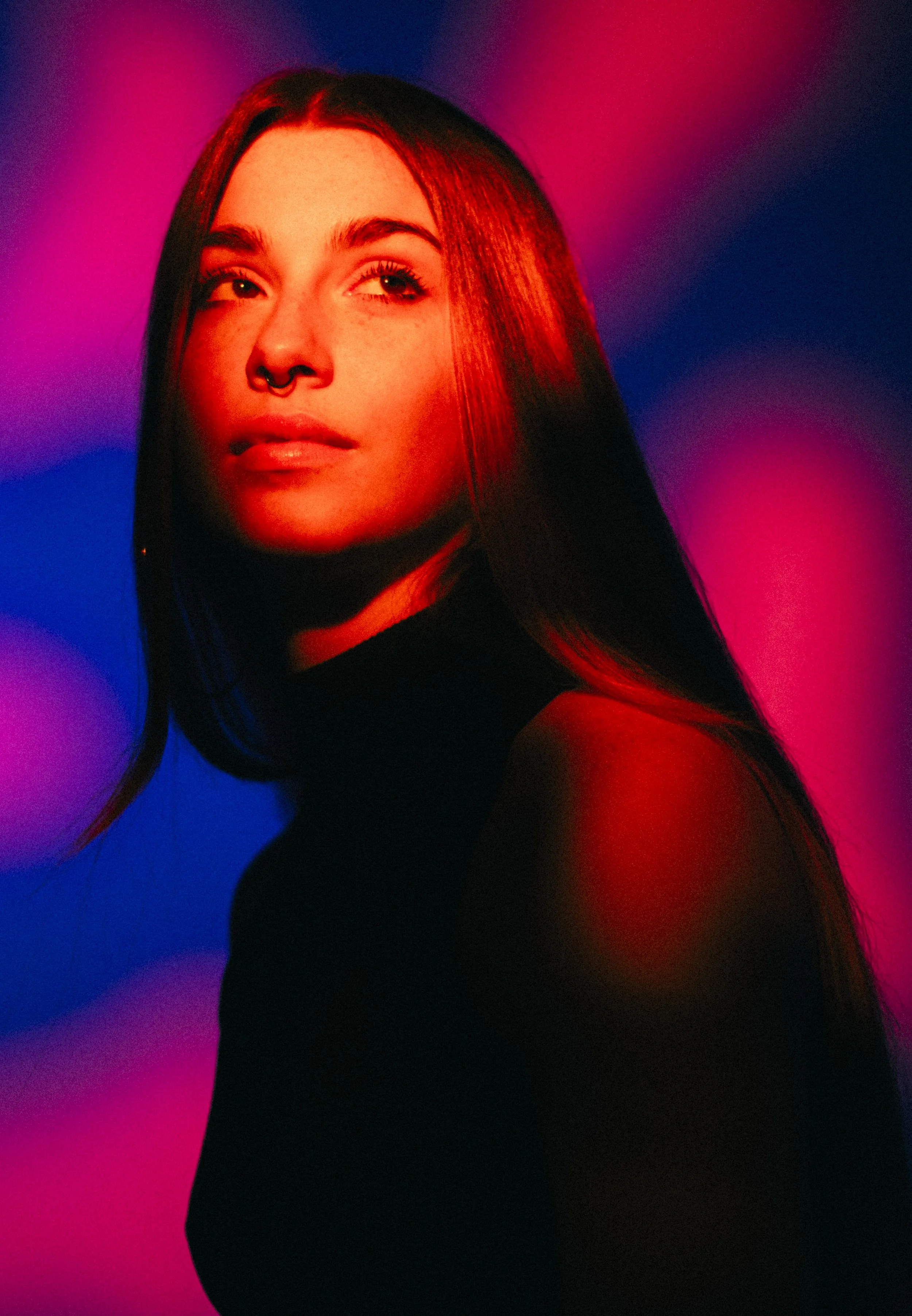 A woman with long dark hair, septum piercing, wearing a black top, illuminated by vibrant pink, purple, and blue lighting.