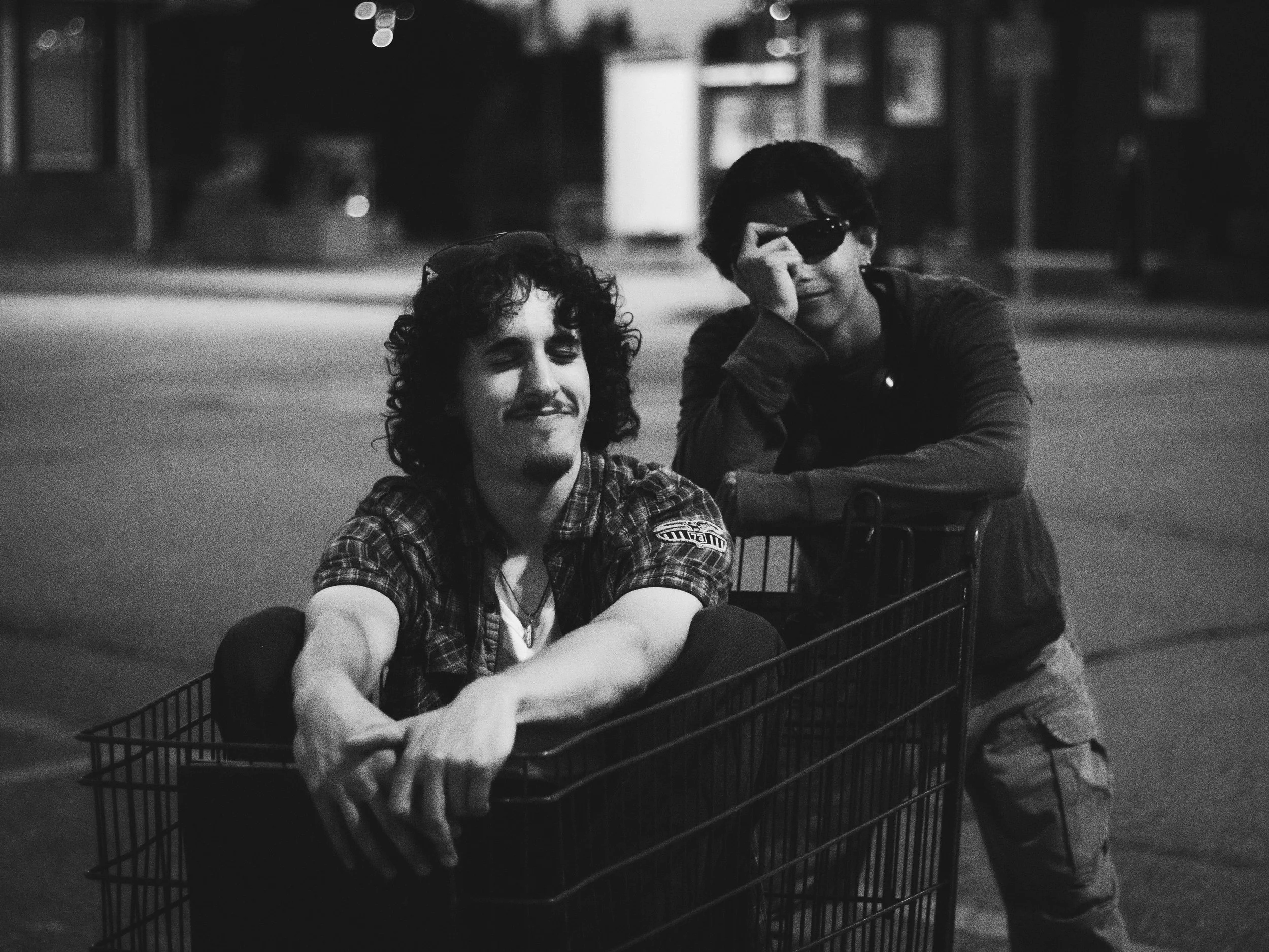 Two young men, one sitting in a shopping cart and the other standing behind it, are outdoors at night. The man in the cart has curly hair, a mustache, and is wearing a plaid shirt, while the man standing behind is wearing sunglasses, a jacket, and ca