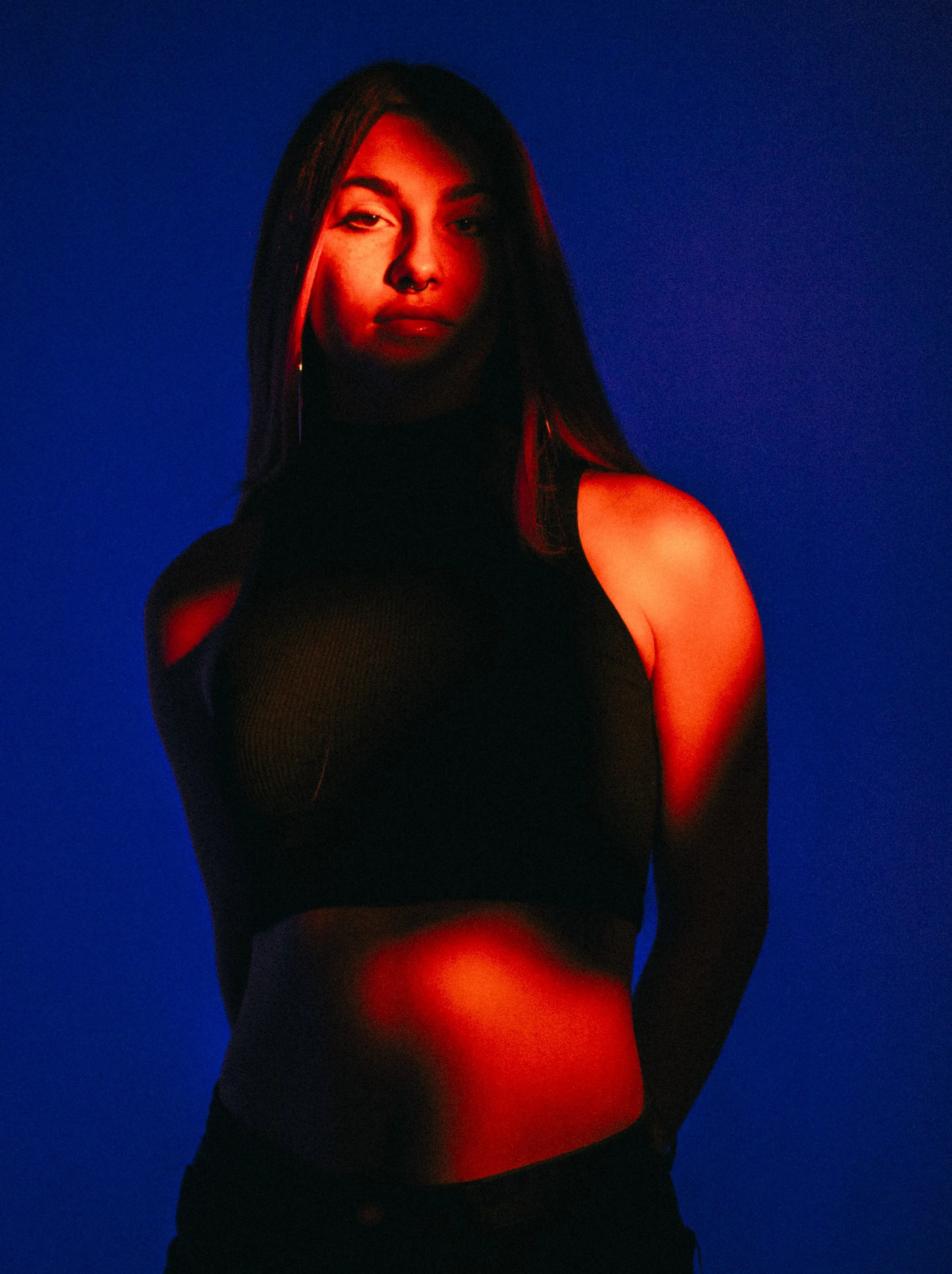 A woman with long dark hair wearing a black sleeveless top standing against a blue background, illuminated by red and blue lighting.