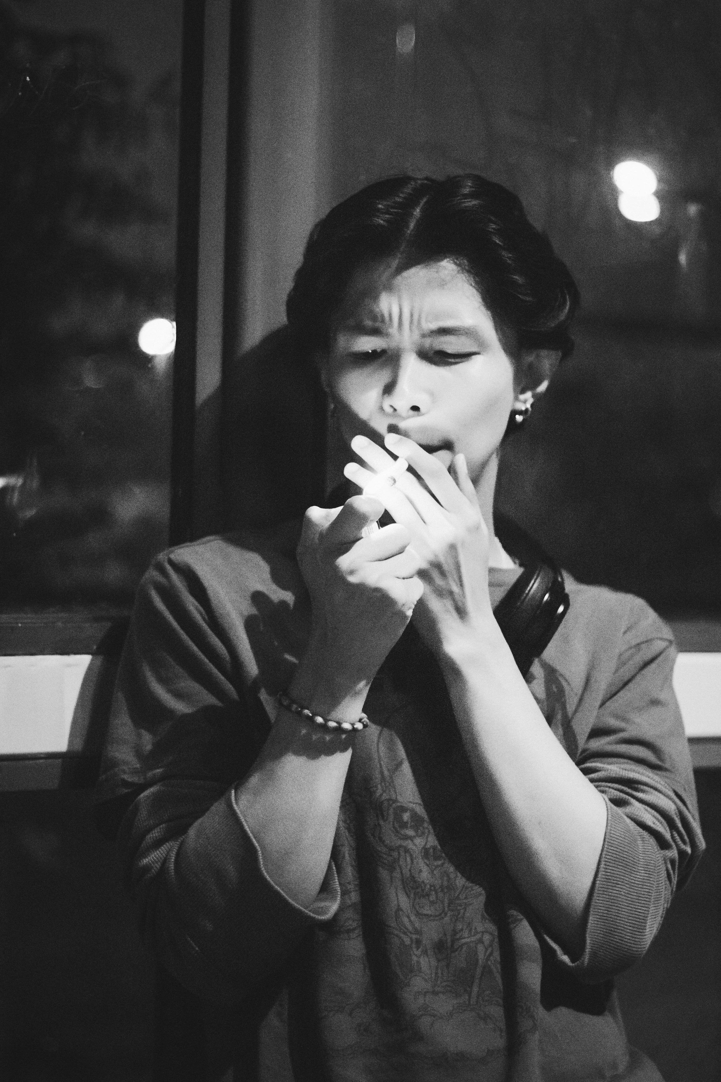 A woman with short dark hair, wearing earrings and a bracelet, is smoking a cigarette while holding it in her hand, with a contemplative expression on her face, in a black-and-white photo.
