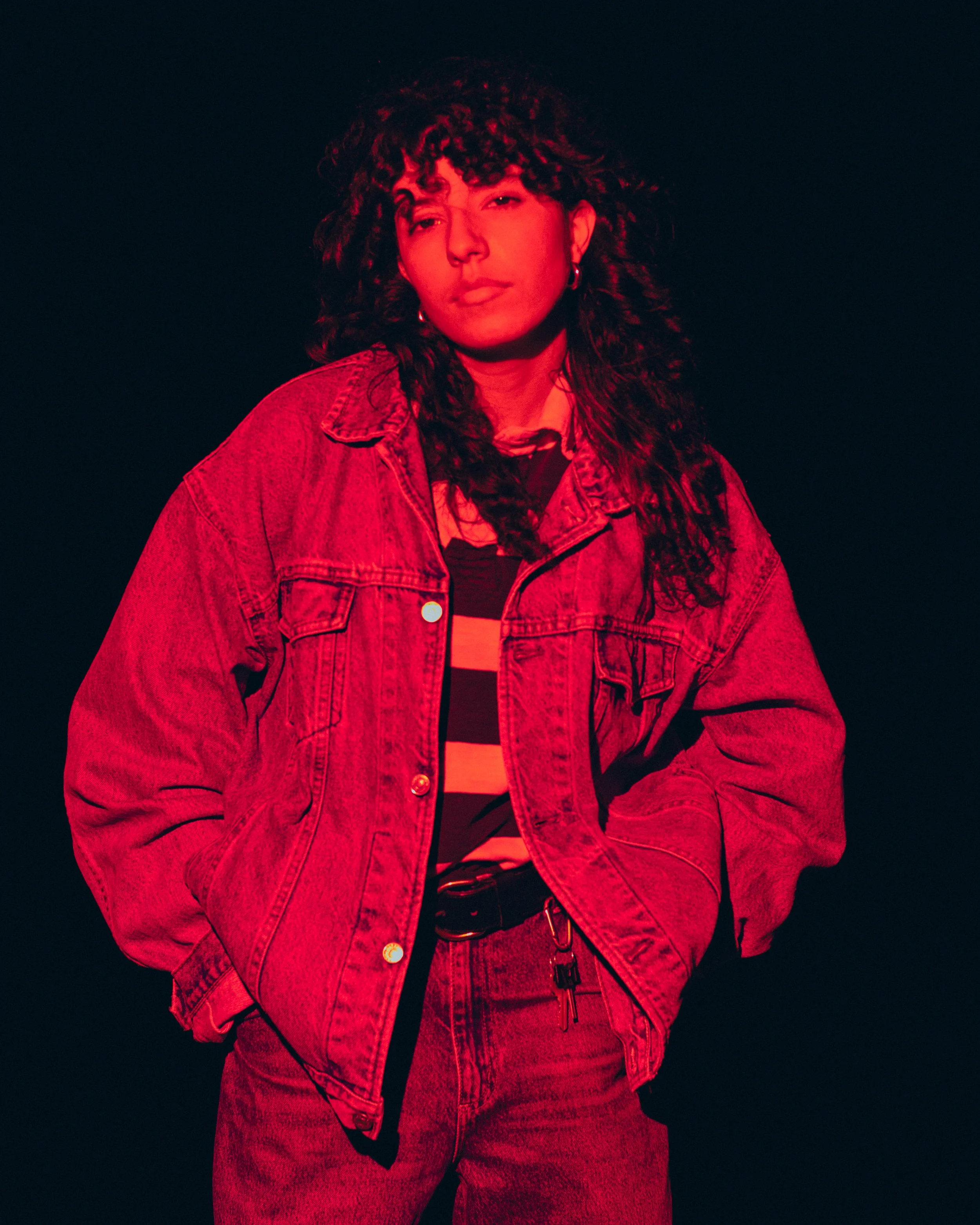 Person with curly hair wearing a denim jacket and striped shirt, standing against a dark background with red lighting.