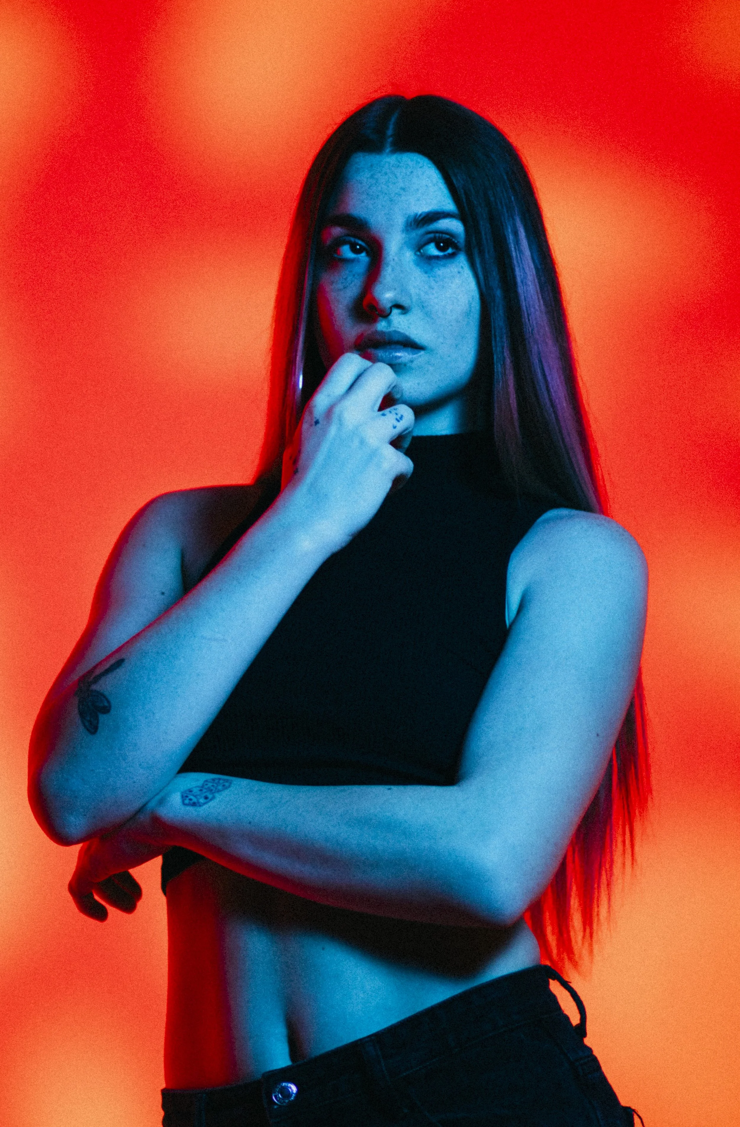 A woman with long dark hair, blue makeup, and tattoos, posing in a black sleeveless top against a red-orange background.