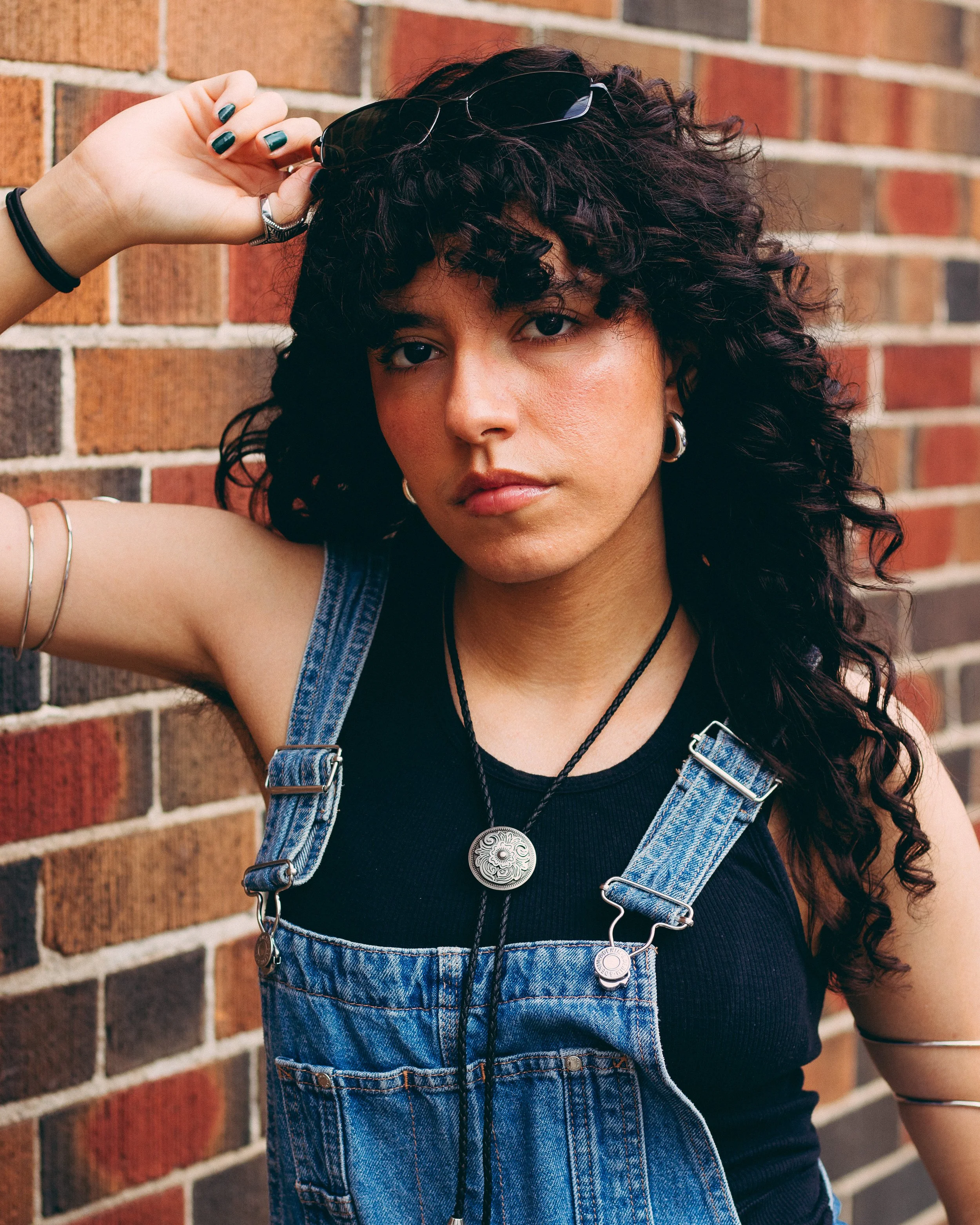 A woman with curly dark hair and hoop earrings, wearing a black sleeveless top and denim overalls, standing against a brick wall, holding sunglasses on her head with her right hand.