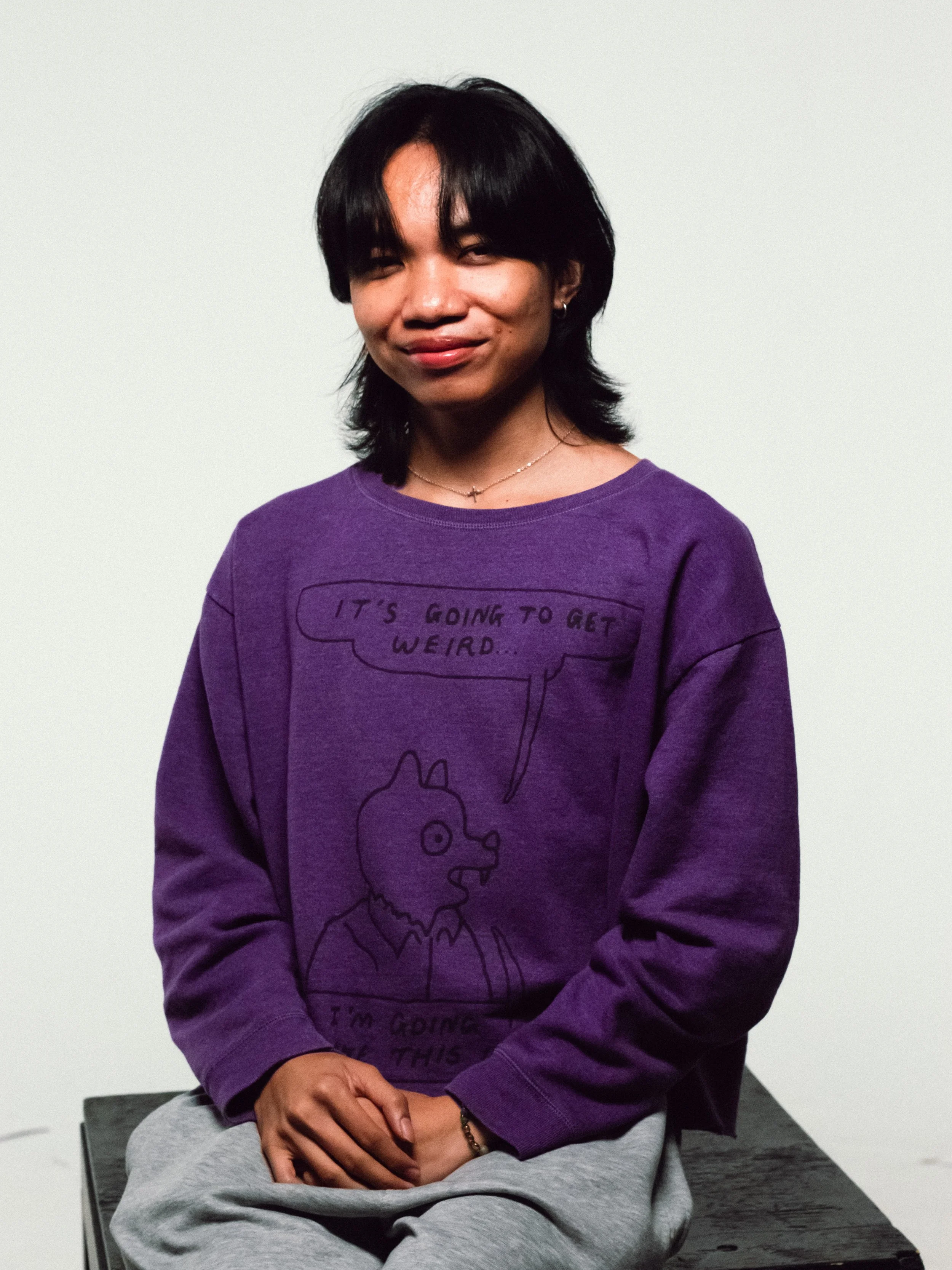 A person with shoulder-length black hair, wearing a purple sweatshirt with a cartoon dog and text, sitting on a black surface against a plain white background.