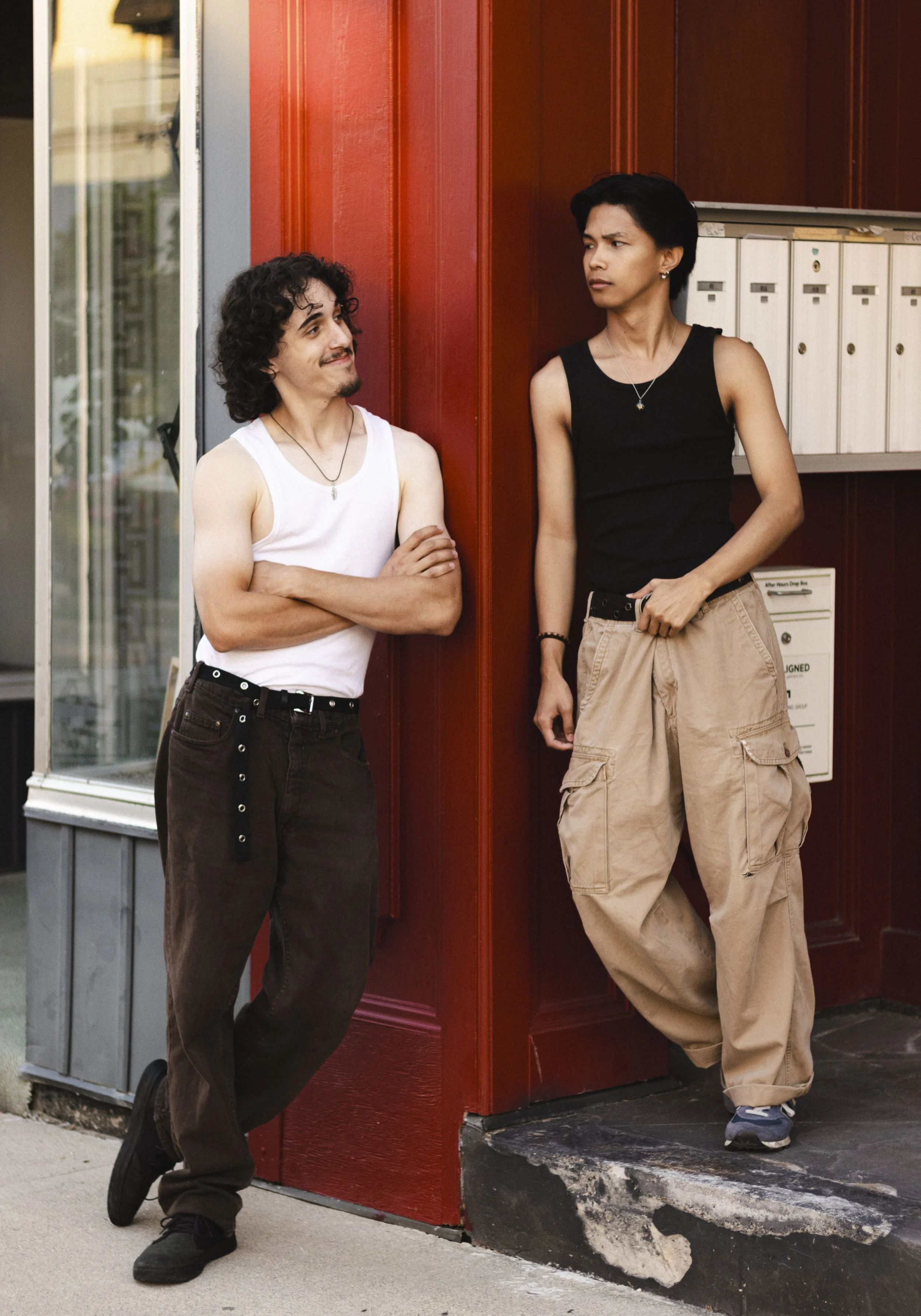 Two young men are standing and leaning against a red wall near corner of a building with mailboxes and glass windowed door. One is wearing a white tank top and dark baggy pants, while the other is wearing a black tank top and tan cargo pants. They ar
