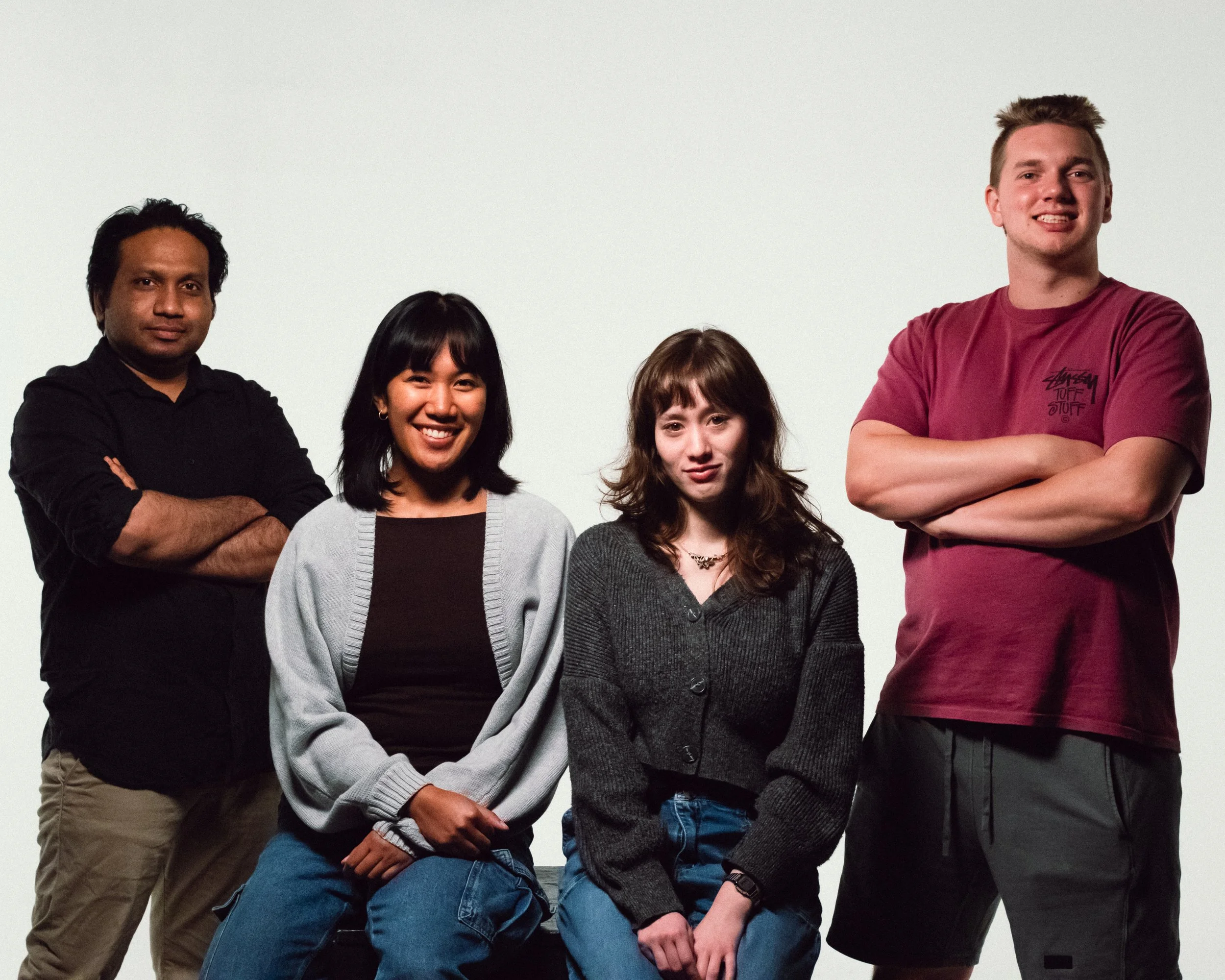 Group of four young adults posing against a plain light background, with two women sitting and two men standing behind them.