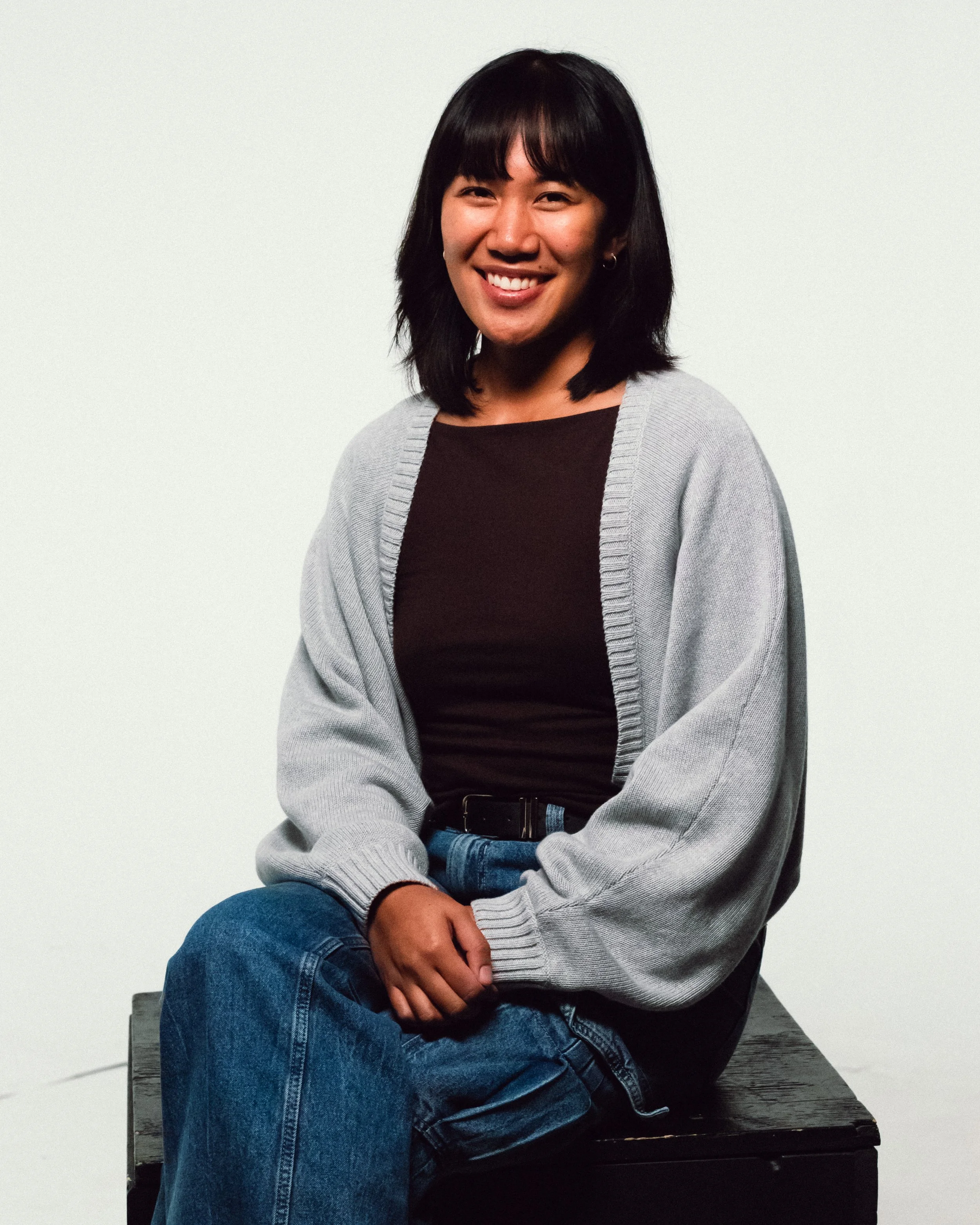 A young woman with black hair and a smile, sitting on a wooden box against a plain white background, wearing a gray cardigan, black top, and blue jeans.