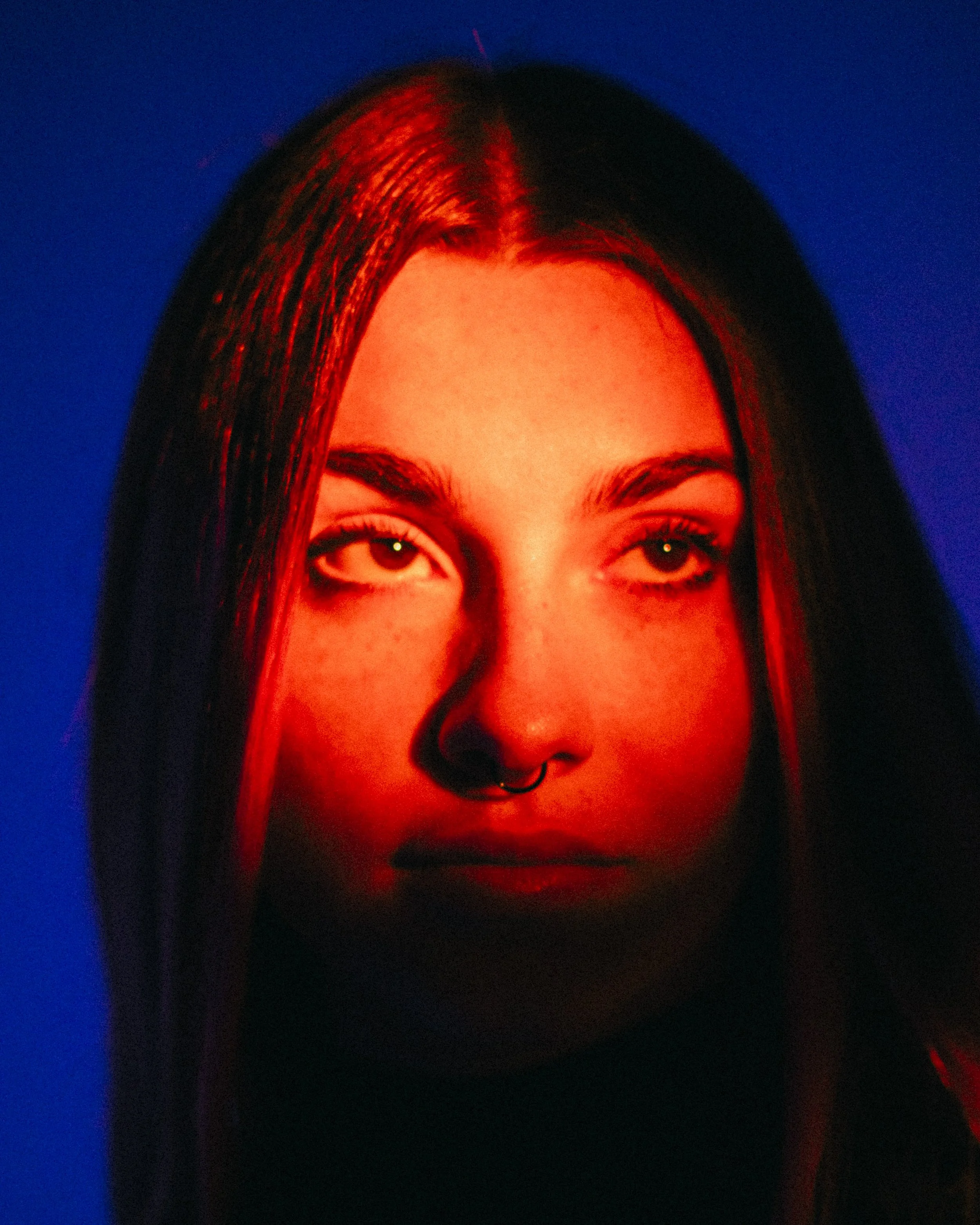 Close-up portrait of a young woman with long dark hair, glowing red lighting on her face, wearing a septum piercing, against a dark background.