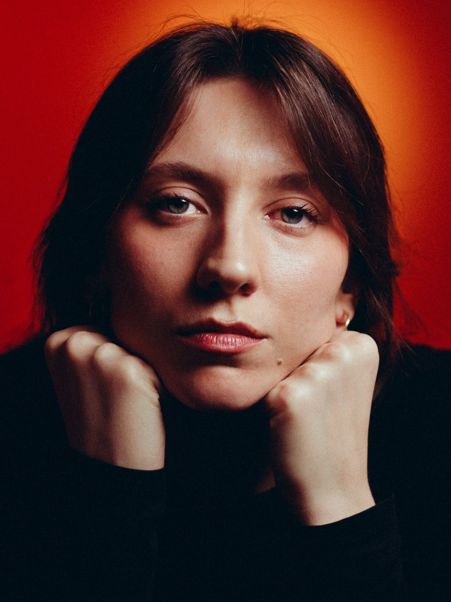 A woman with fair skin and short dark hair, resting her chin on her hands, against a warm red and orange background.
