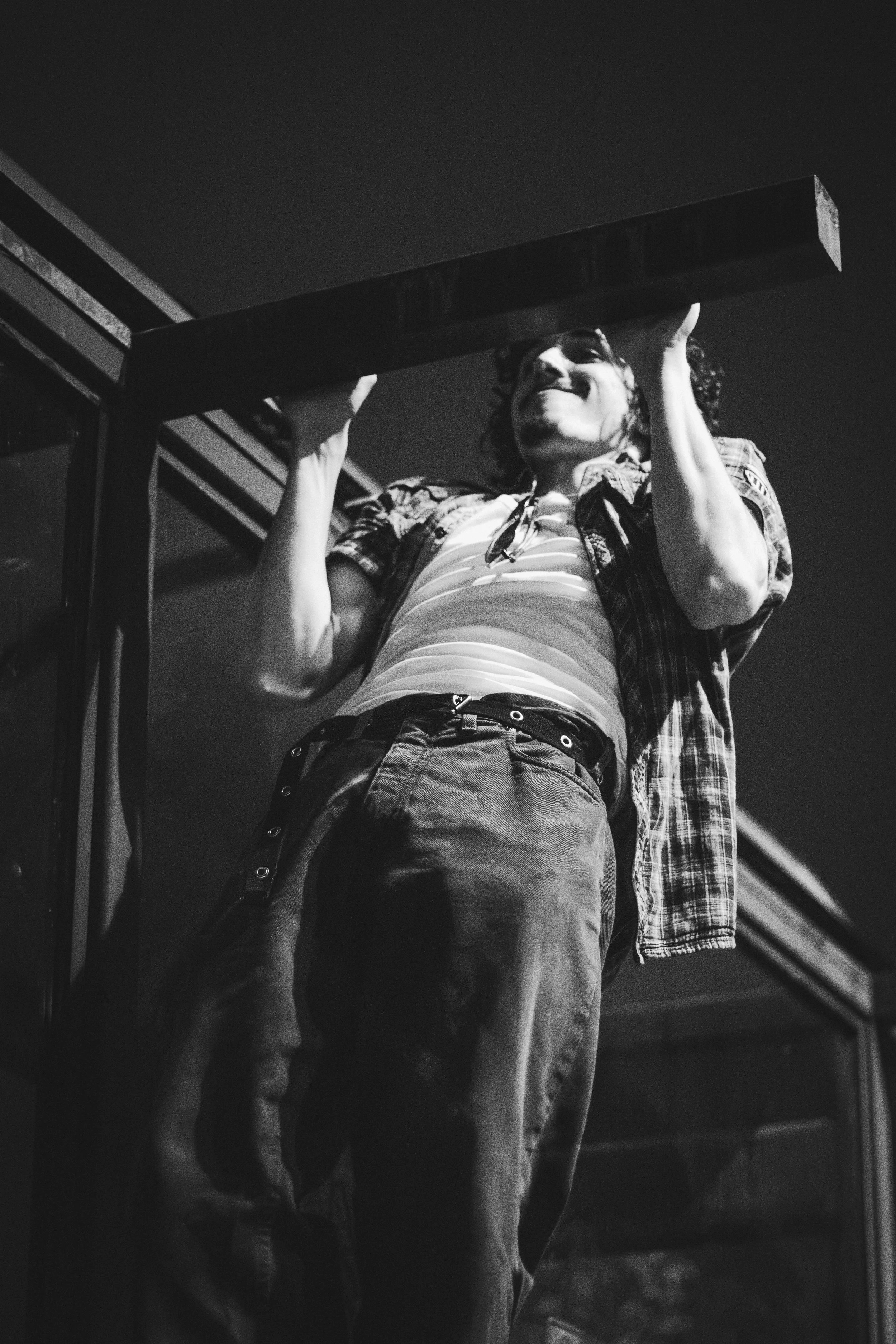 A person with curly hair smiling while lifting a large wooden plank overhead, standing outdoors at night.