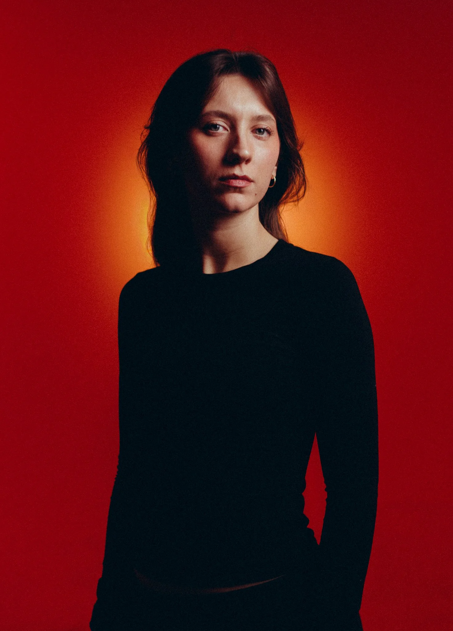 Portrait of a young woman with shoulder-length brown hair wearing a black top, standing against a red and orange gradient background.