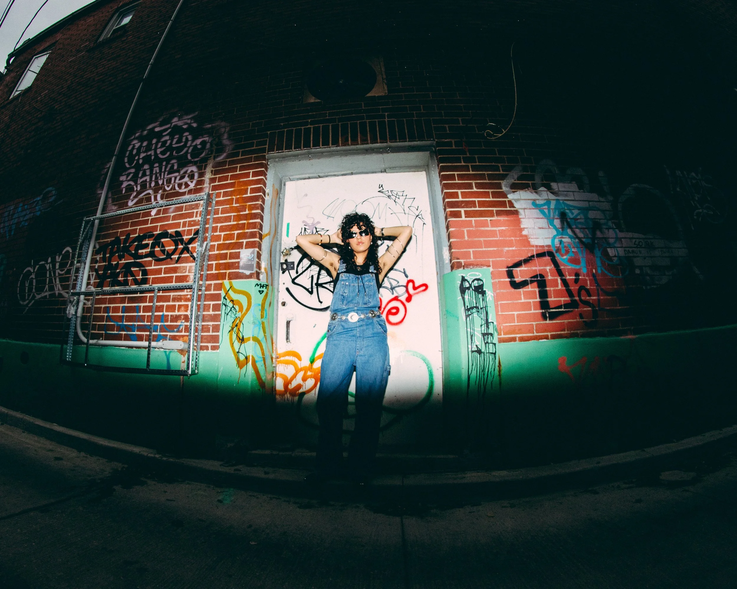 A woman with dark curly hair, sunglasses, wearing overalls, standing in front of a graffiti-covered brick wall