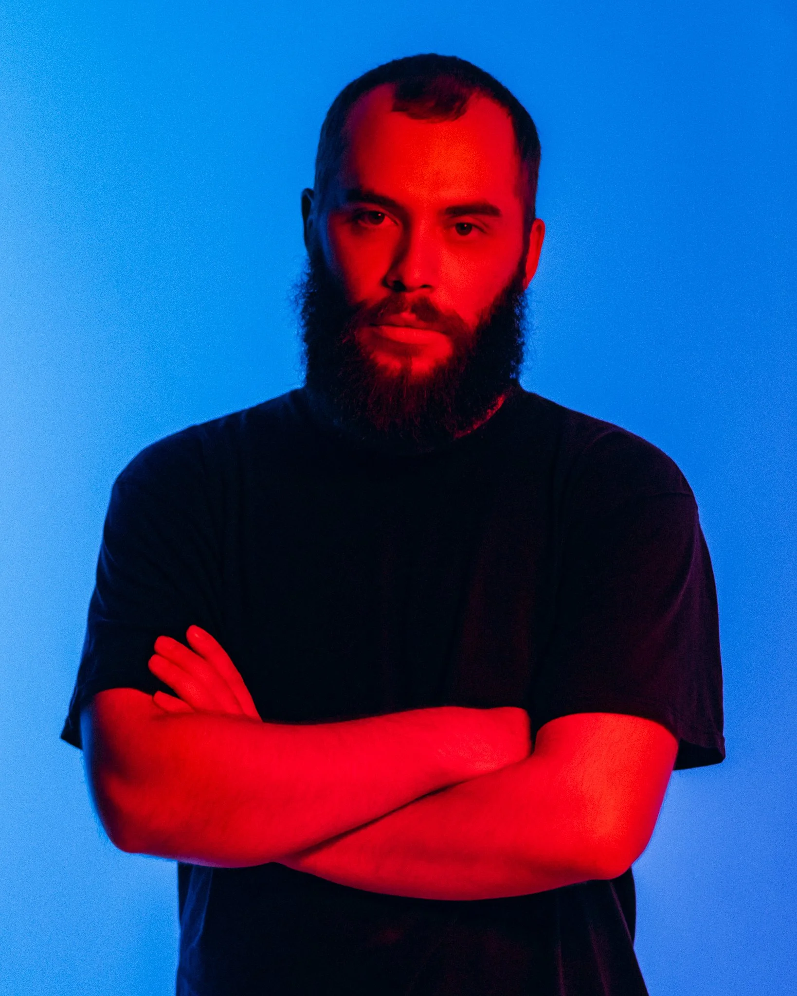 A man with a beard and short hair, wearing a black t-shirt, standing with arms crossed against a blue background with red lighting.