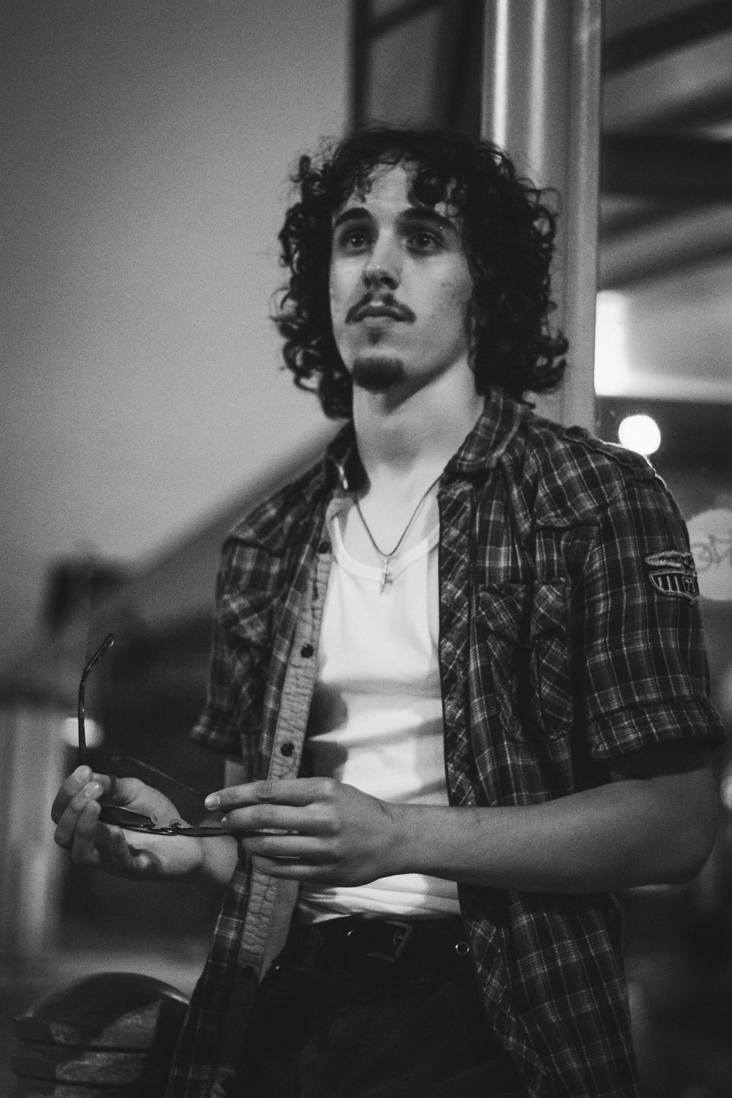 A young man with curly hair, mustache, and goatee wearing a checkered shirt over a white t-shirt holding glasses, standing indoors.