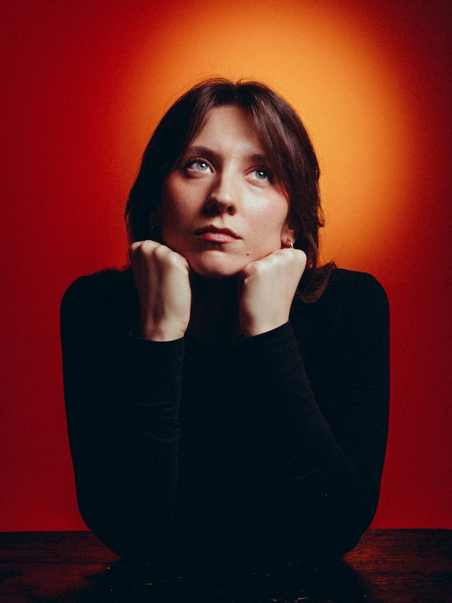 A woman with dark hair resting her chin on her fists, looking upwards against a red and orange background.