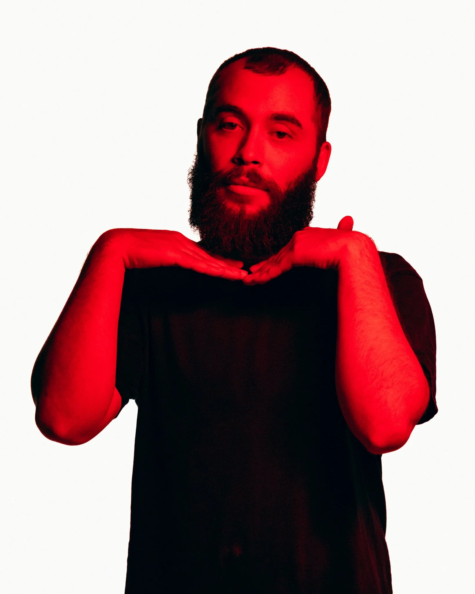 Man with beard in black shirt making heart shape with hands, red lighting on face, white background