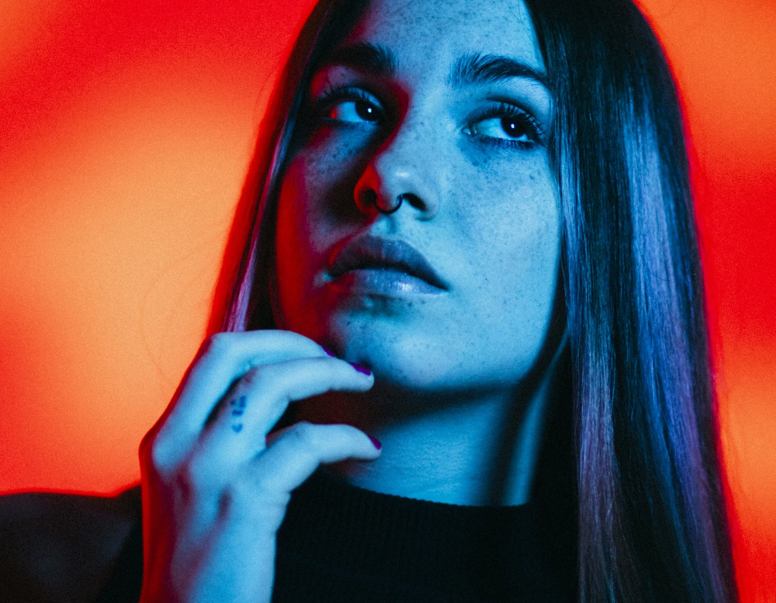 A young woman with long straight hair, wearing a black top, gazing off to the side, illuminated by blue and orange lighting.